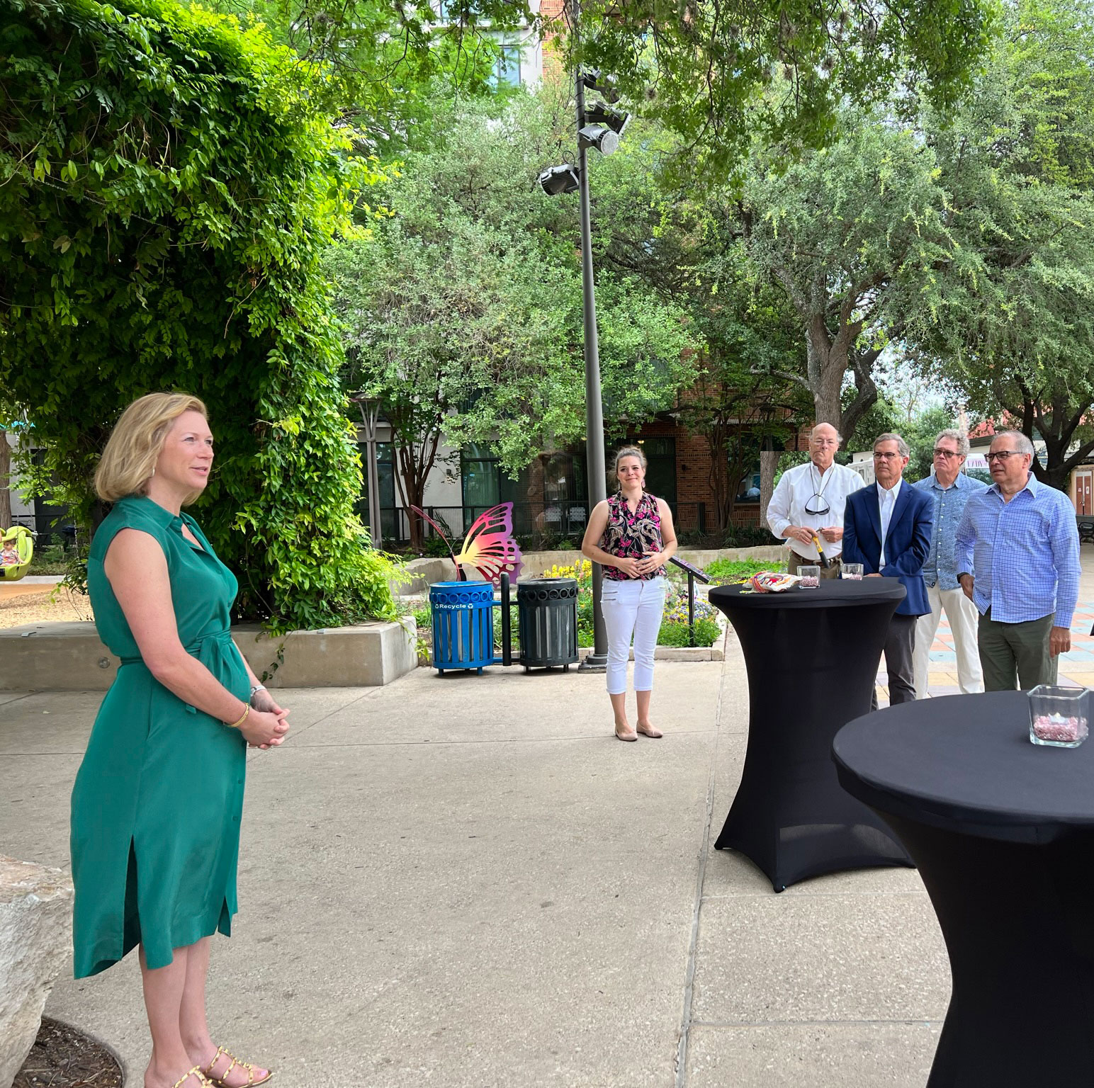 A woman in a green dress speaks to a small group of people outdoors by a table with a black tablecloth, celebrating Arbor Day. Trees and greenery are in the background.
