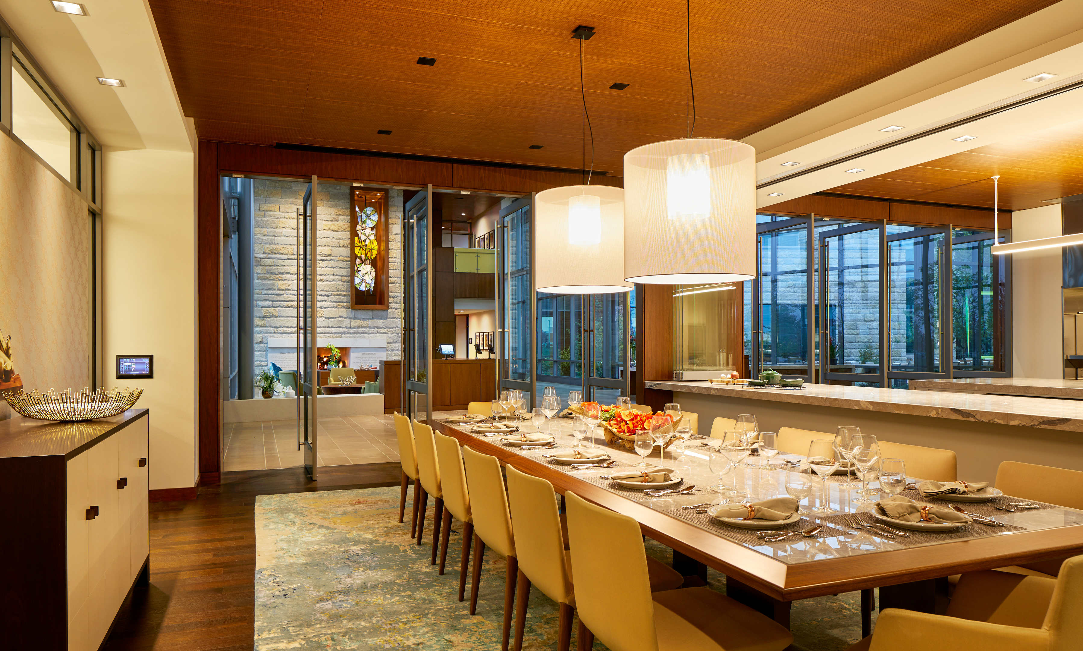 A modern dining room with a long wooden table set for ten, beige chairs, contemporary lighting fixtures, and large windows offering a view into another stylish, open-plan home office.