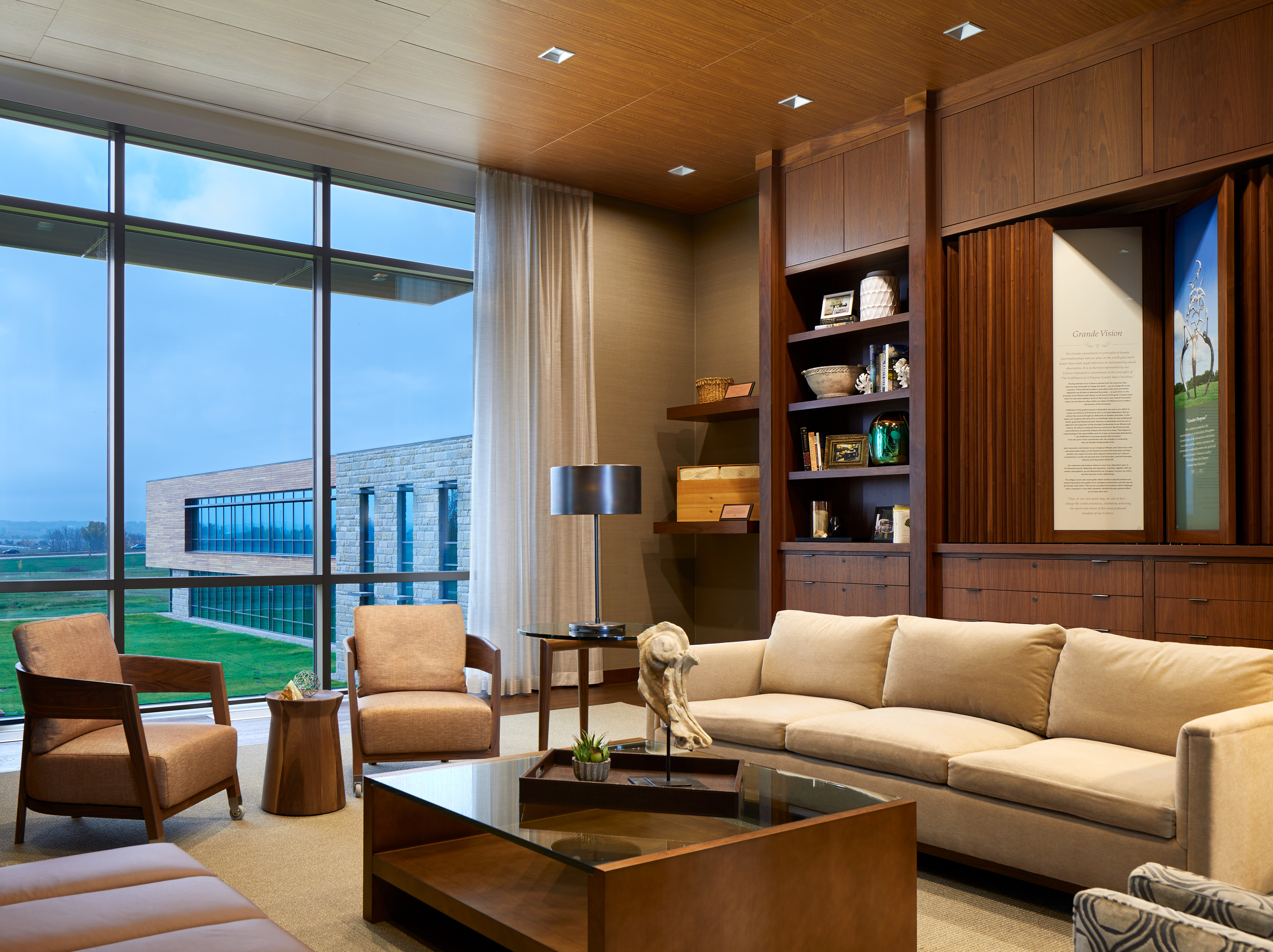 A modern, well-lit lounge area featuring a beige sectional sofa, armchairs, a glass-top coffee table, and built-in wooden shelves. Large windows reveal a view of a contemporary research center and green lawn outside.