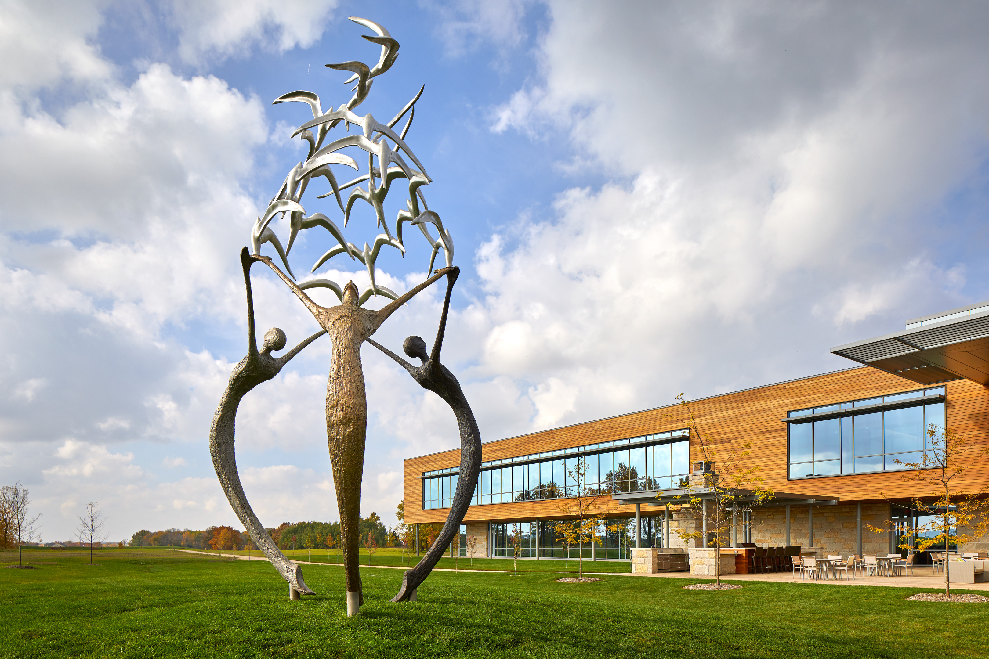 A large outdoor sculpture of three human figures reaching upward, holding bird-like forms, stands prominently in front of a modern research center with expansive windows. The sky is partly cloudy, creating a dramatic backdrop for the art piece.