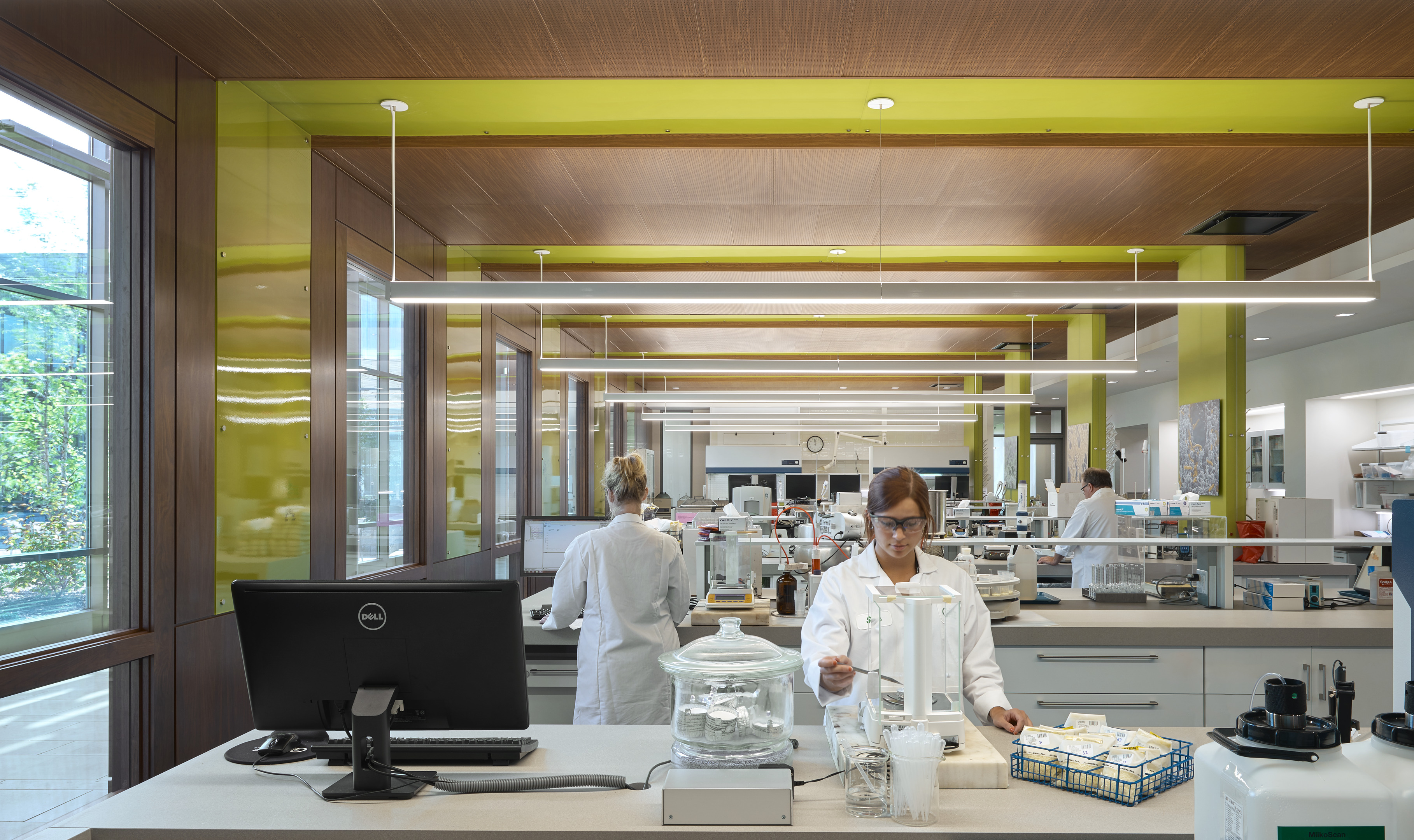 Scientists wearing lab coats work in a modern research center laboratory with various equipment and supplies around them. One scientist is handling a piece of equipment while others work at their stations in the background.