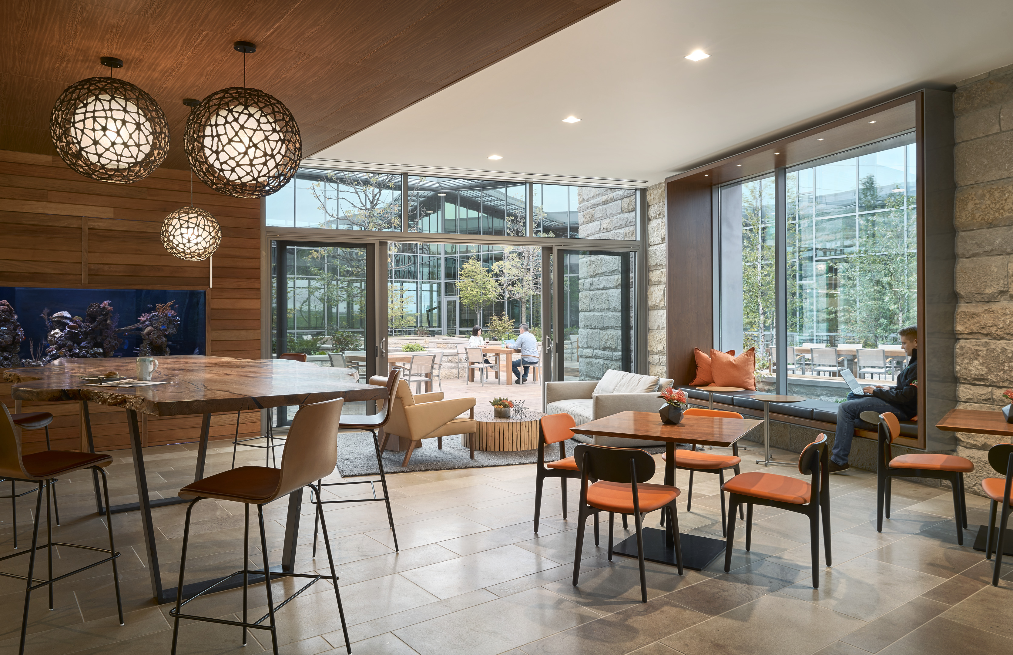 Modern indoor lounge area with wooden tables, colorful chairs, and hanging spherical light fixtures. Large windows reveal an outdoor patio with seating and greenery. One person sits by a window, perhaps transforming the space into a cozy home office for the afternoon.
