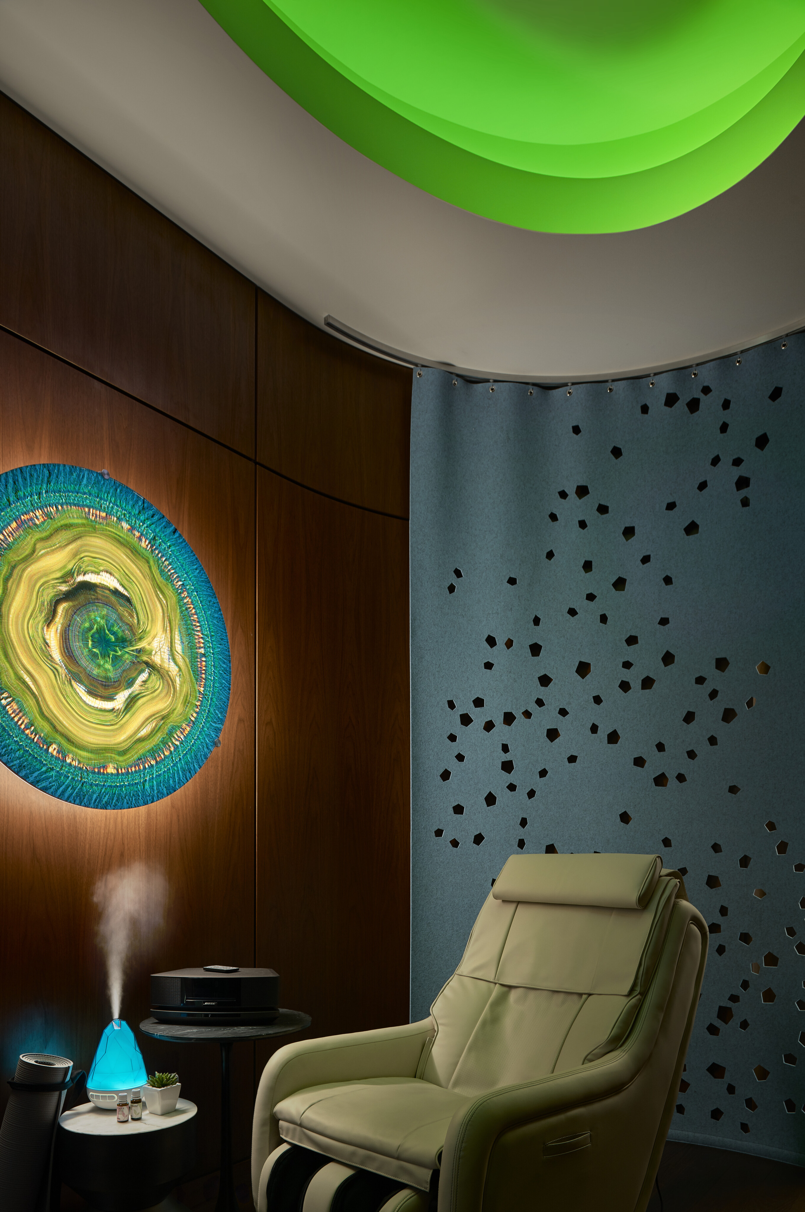A modern relaxation room in the home office features a reclining chair, green ambient ceiling light, abstract wall art, a blue humidifier emitting steam, a small table with a white cup, and a black electronic device.