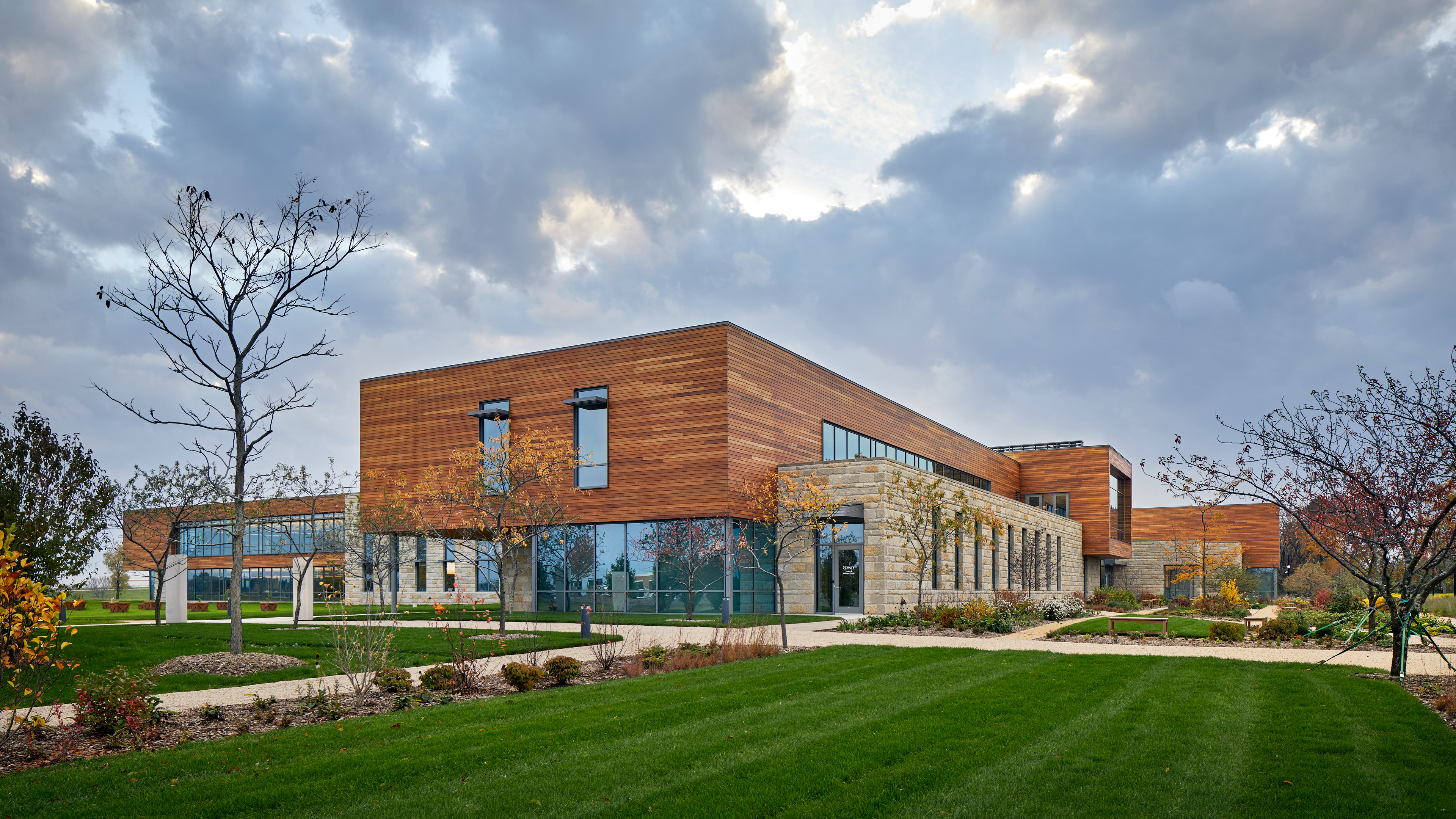 Modern two-story building with wood and stone exterior, surrounded by green lawn and paths under a cloudy sky. Sparse trees and landscaped garden areas are visible in the foreground, giving it the serene atmosphere of a Research Center.