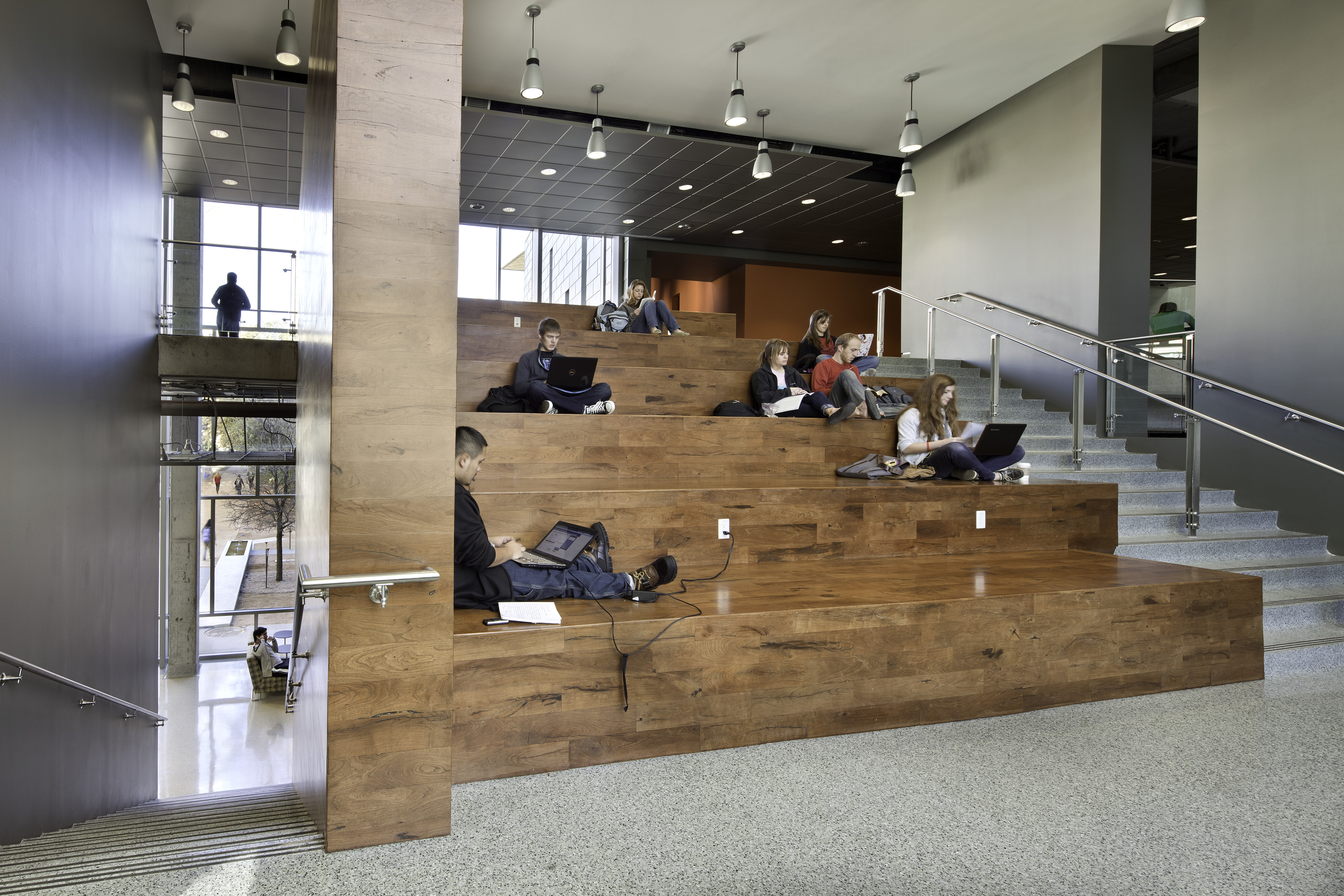 Several people sit on large wooden steps in a modern indoor space at the University of Texas Student Activity Center, working on laptops or reading, with others visible in the background.
