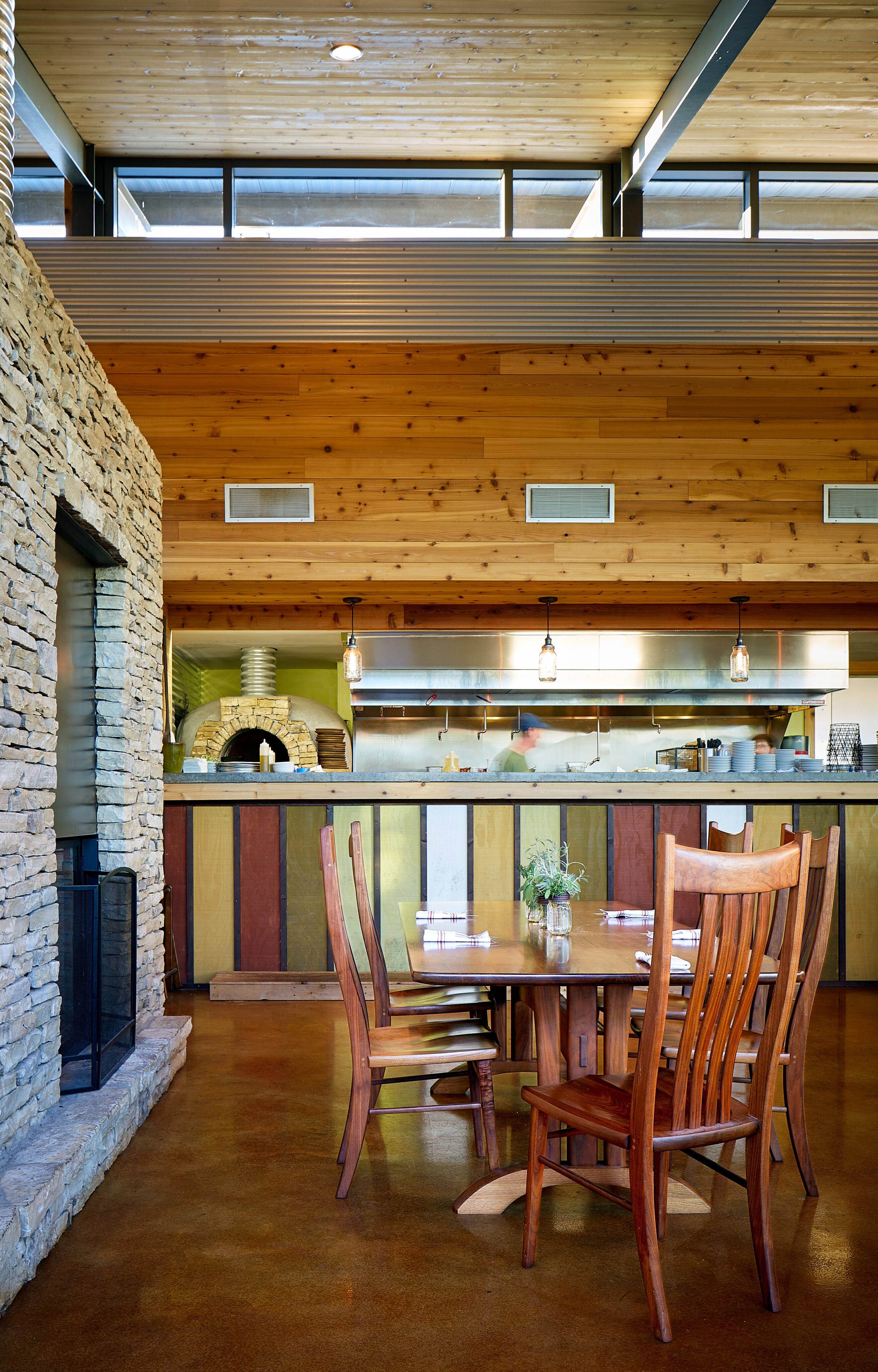 A rustic dining area features a wooden table with four chairs, a stone wall, and a kitchen with a pizza oven in the background. Pendant lights hang over the counter, adding charm to this cozy space highlighted by important keywords for homey comfort.