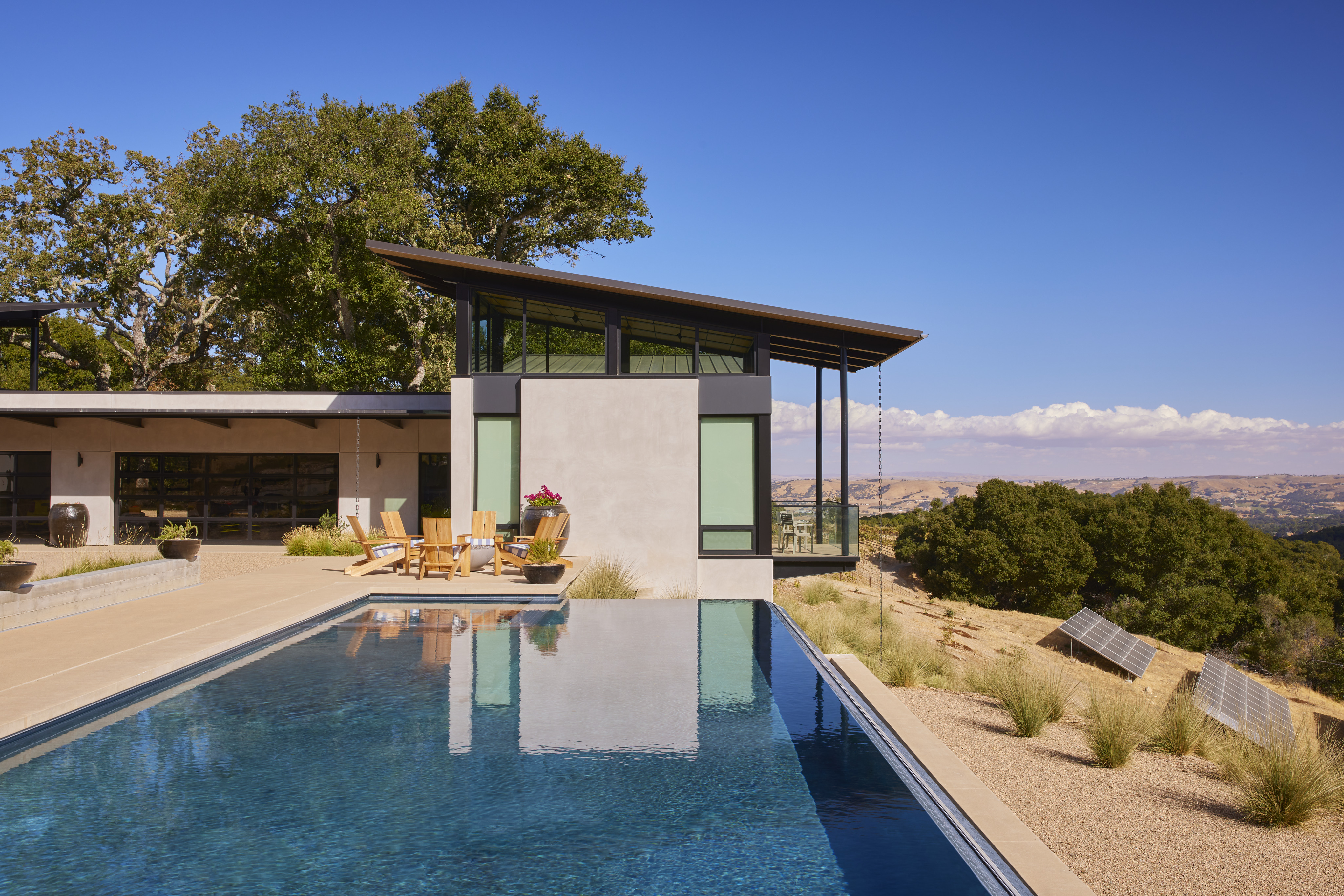Modern house with large windows beside a swimming pool at MoonSprings Vineyard, surrounded by trees and hills under a clear blue sky. Several wooden chairs are arranged on the patio.