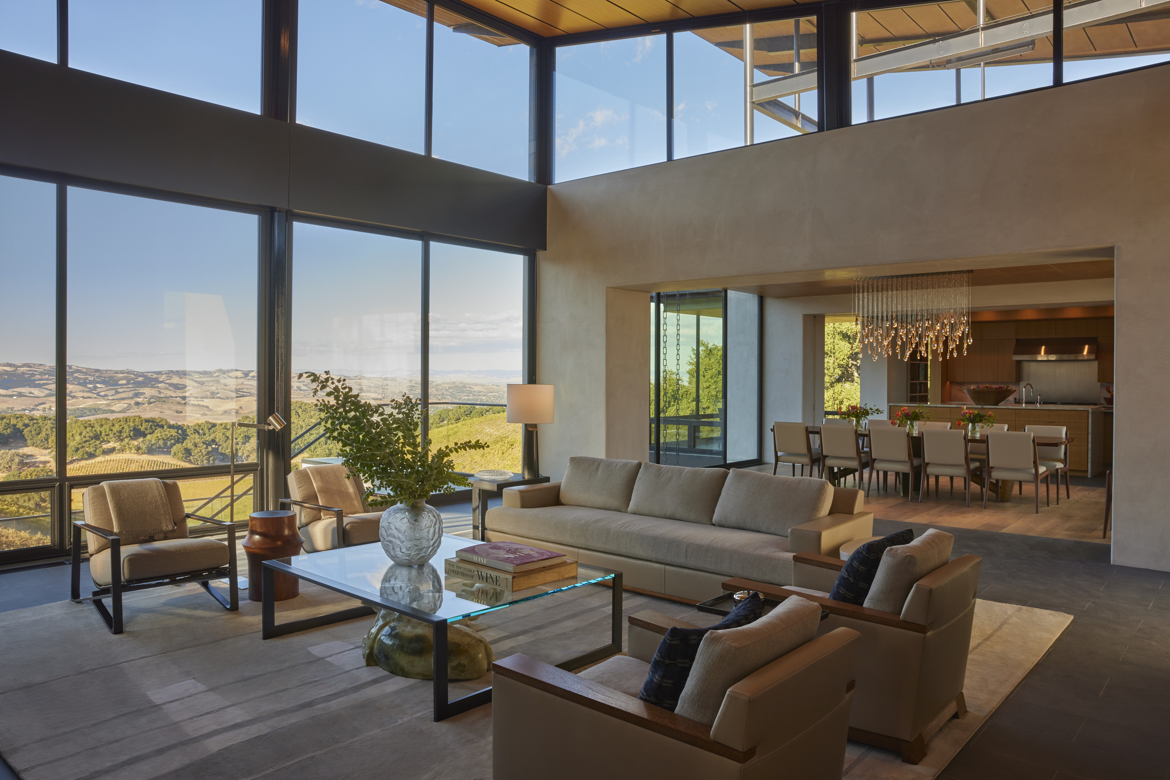Modern open-concept living and dining area with large floor-to-ceiling windows, neutral-toned furniture, and a scenic view of the rolling hills of MoonSprings Vineyard outside.
