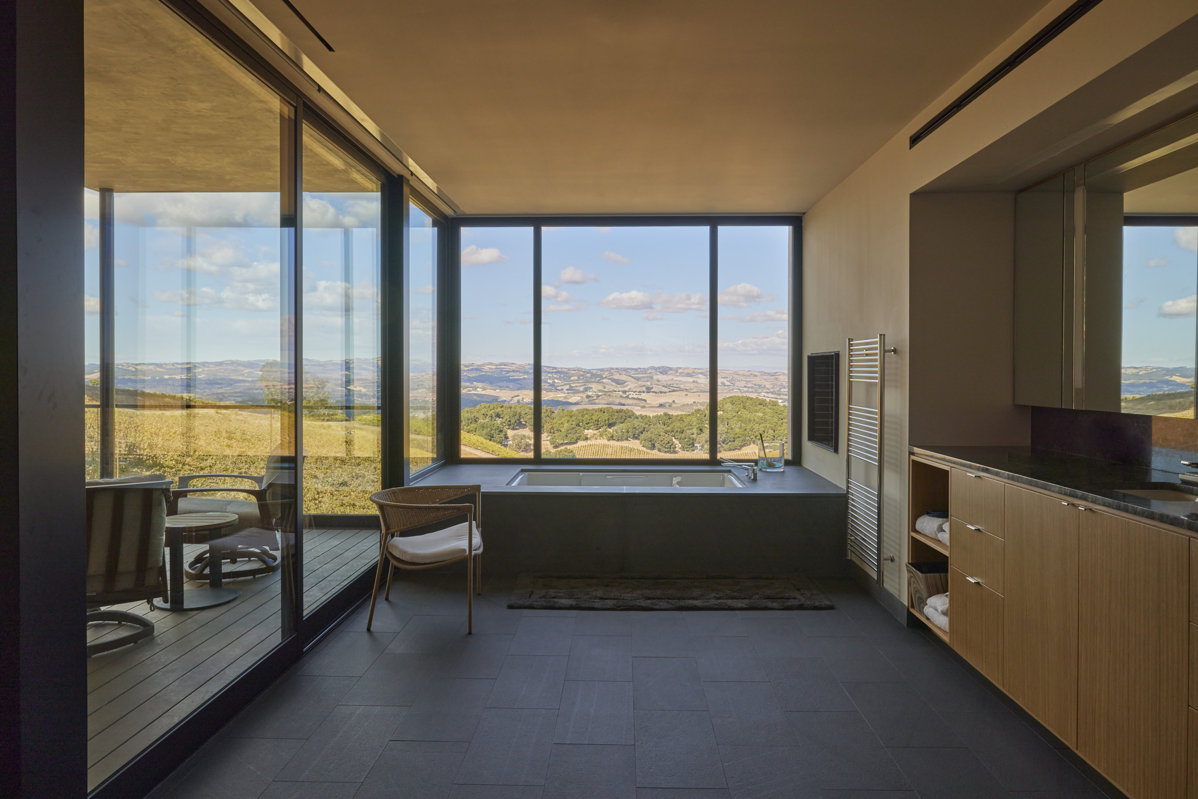 Modern bathroom with large windows, a freestanding bathtub, a chair, open shelving, and an outdoor seating area overlooking the scenic hills and fields of MoonSprings Vineyard.
