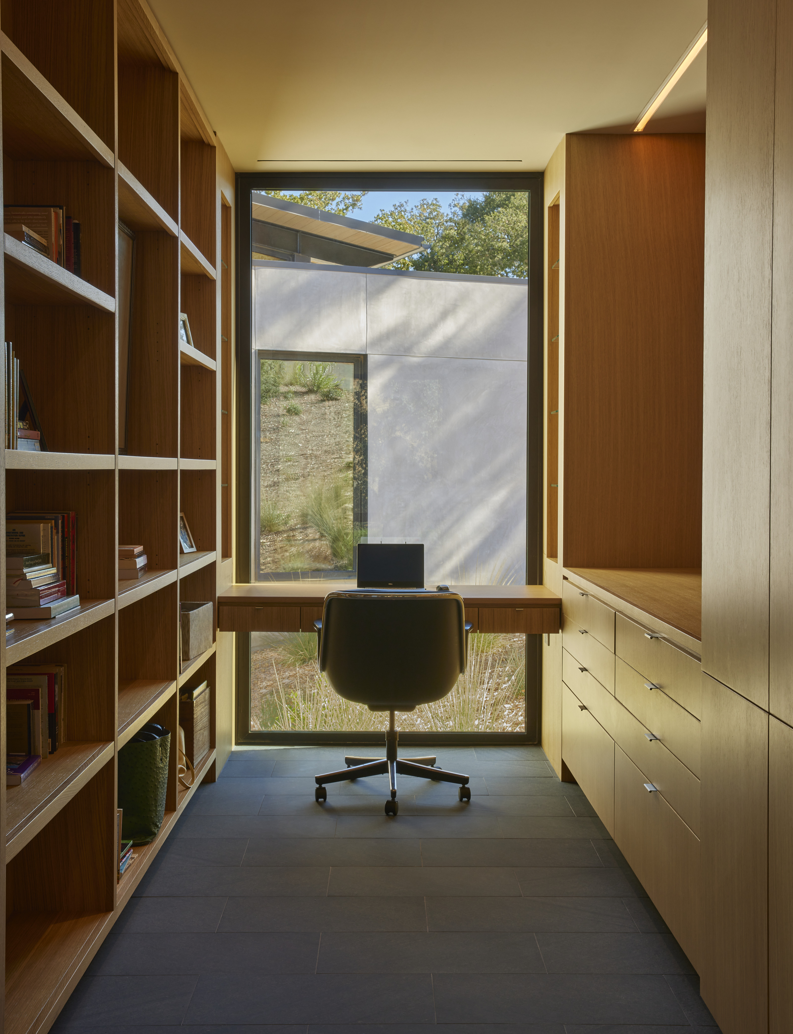 A modern home office with wooden shelves and cabinets, a desk facing a large window overlooking MoonSprings Vineyard, a laptop on the desk, and an office chair.