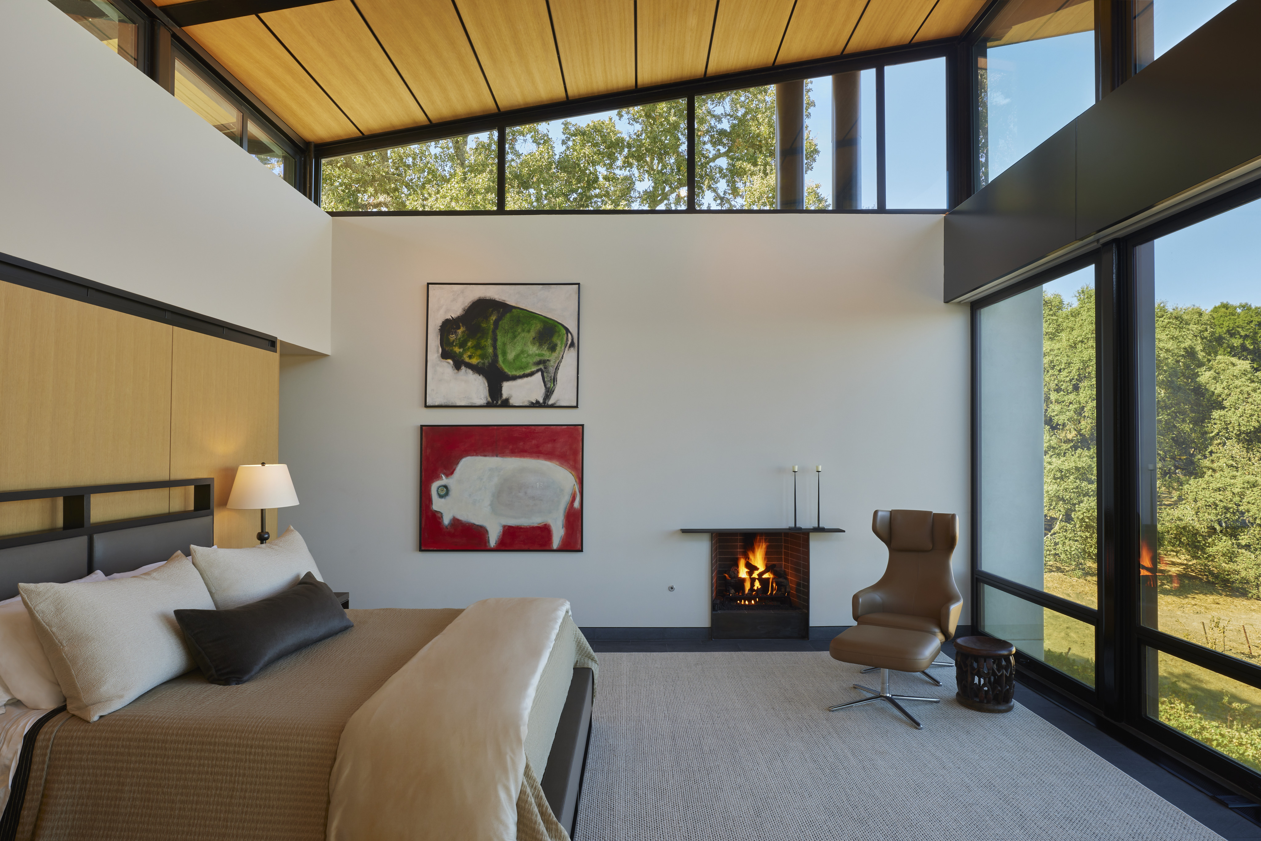Modern bedroom with floor-to-ceiling windows, a bed, and an armchair by a small fireplace. Two animal paintings adorn the wall, while trees from MoonSprings Vineyard are visible just outside.