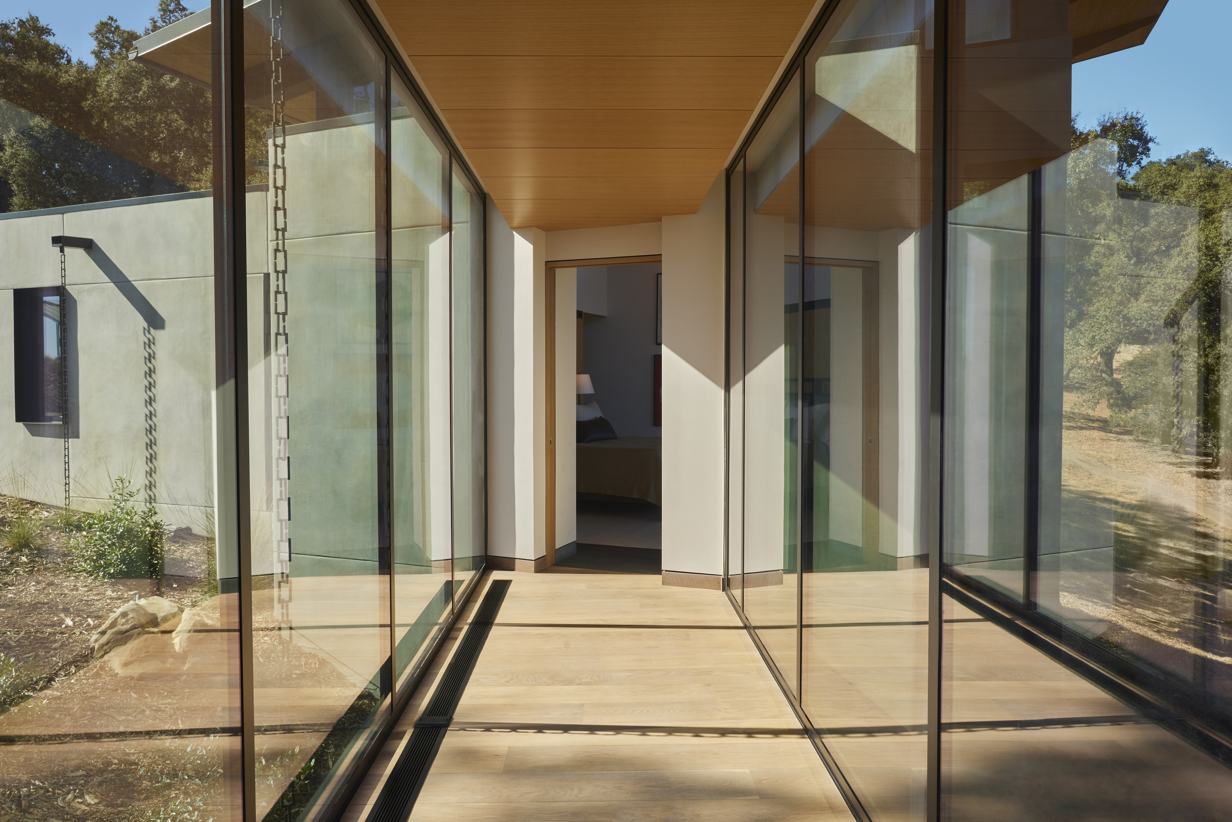 A hallway with floor-to-ceiling glass walls on both sides connects two modern building sections with wood and concrete finishes, offering stunning views of MoonSprings Vineyard.