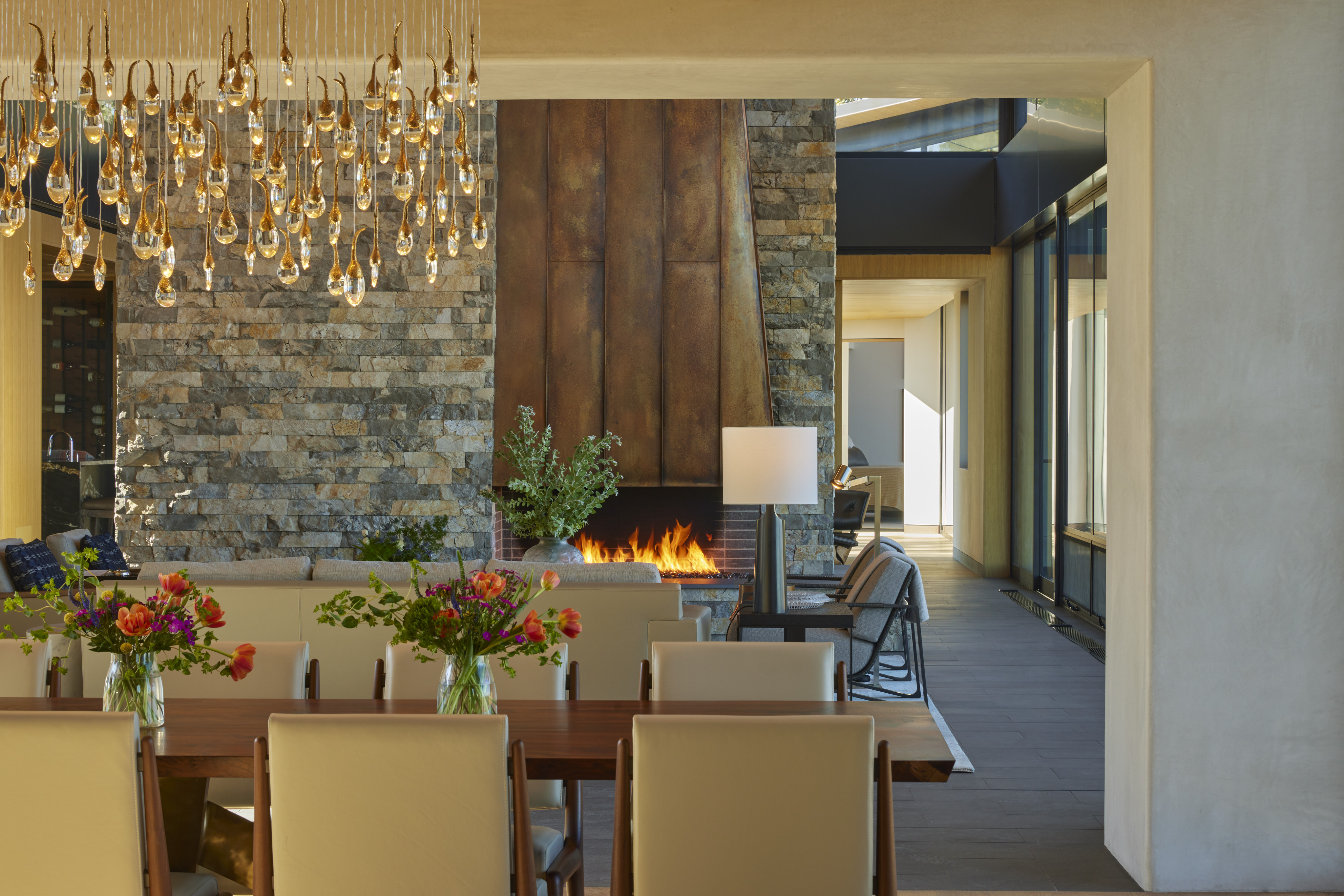 Modern dining room with a wooden table, white chairs, floral centerpieces, stone fireplace, large chandelier, and a view into a contemporary hallway—perfect for gatherings after a day at MoonSprings Vineyard.