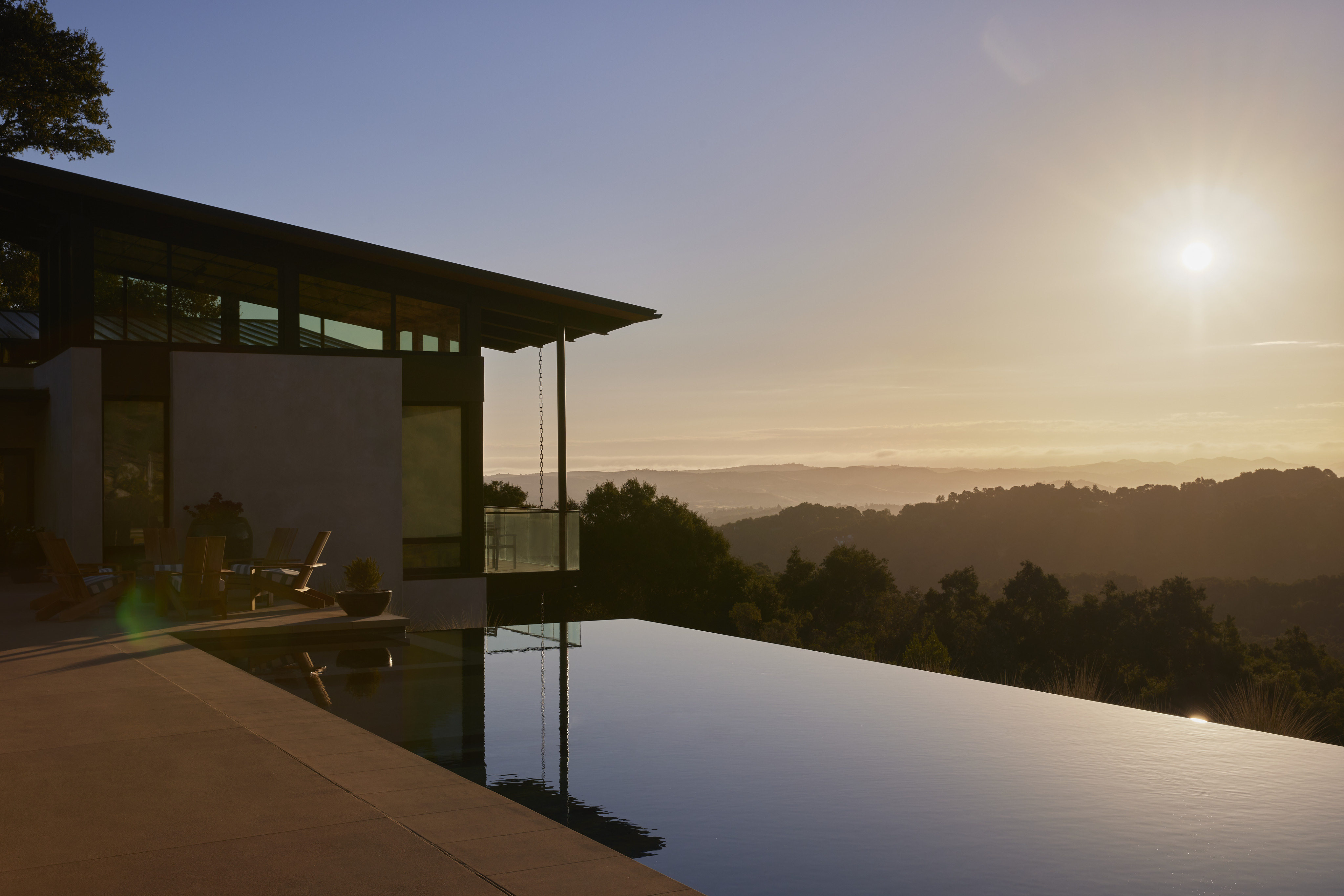 A modern house with a patio overlooks an infinity pool and the scenic MoonSprings Vineyard valley at sunset, with trees and hills in the background.