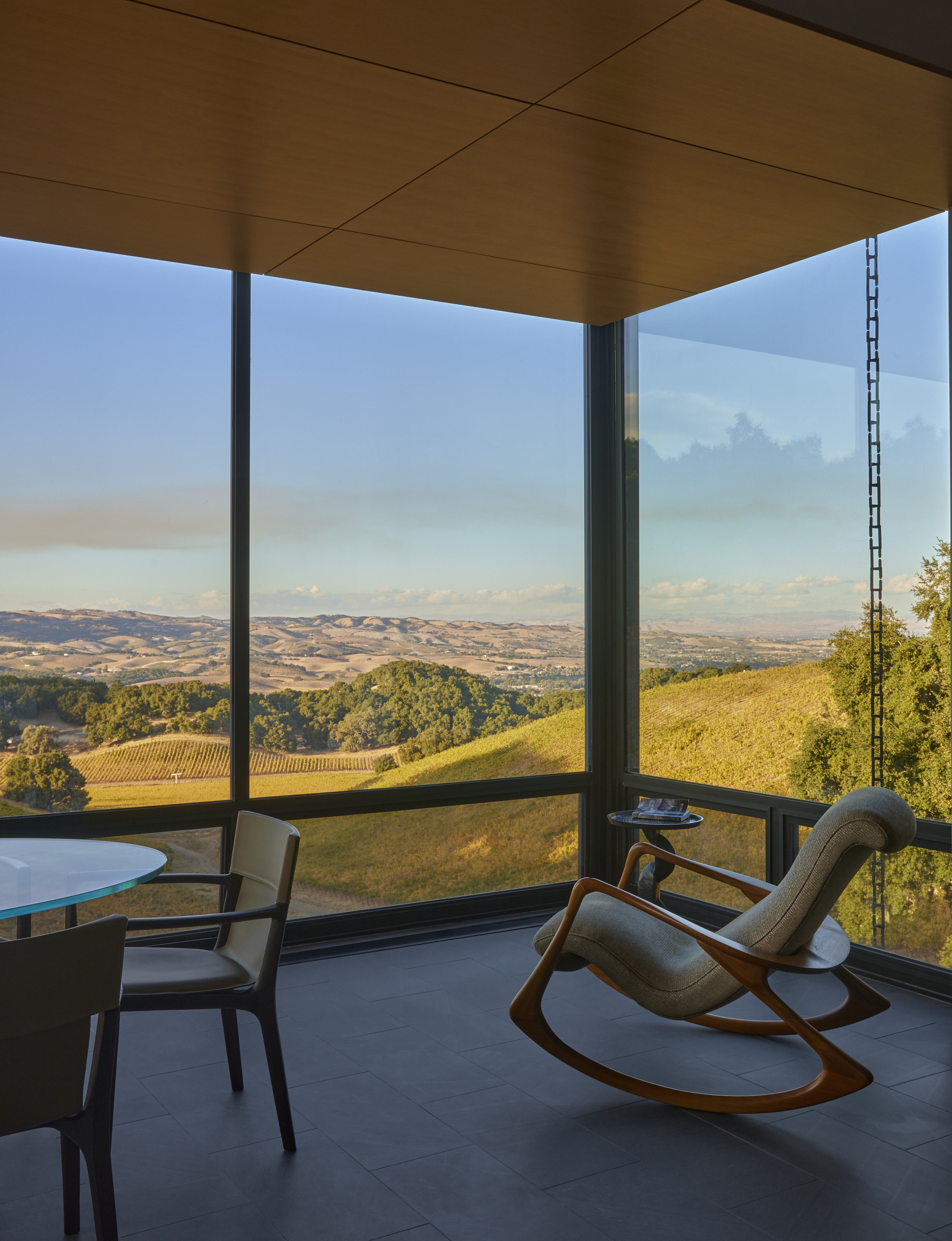 Modern interior with a rocking chair and dining table near large floor-to-ceiling windows overlooking rolling hills and distant mountains under a clear sky, inspired by the serene landscapes of MoonSprings Vineyard.