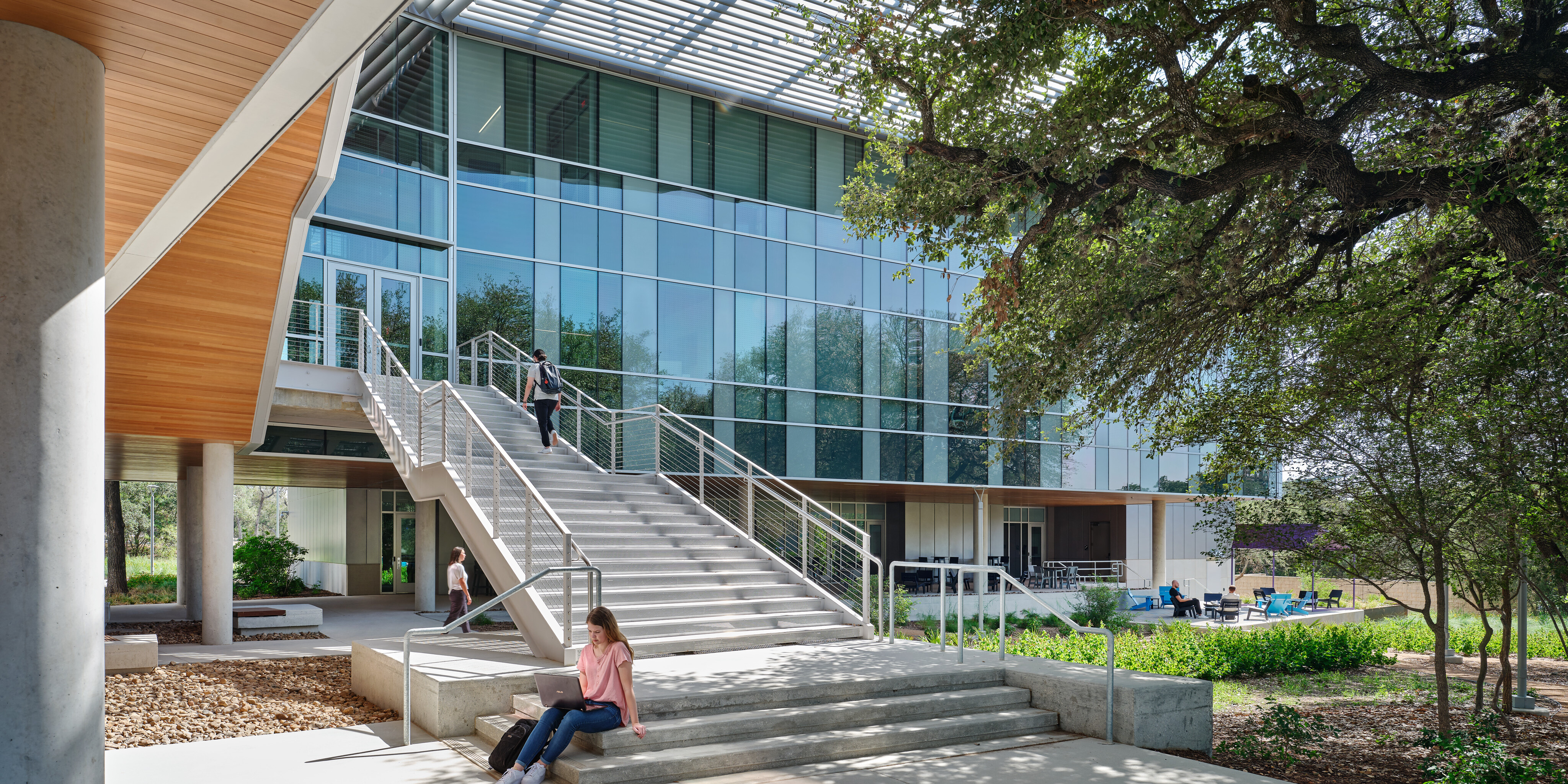 Modern building with a glass façade and outdoor staircase at Northwest Vista College. People sit and walk on the steps, and large trees provide shade. The STEM Center inside fosters innovation and collaboration among students from Alamo Colleges.