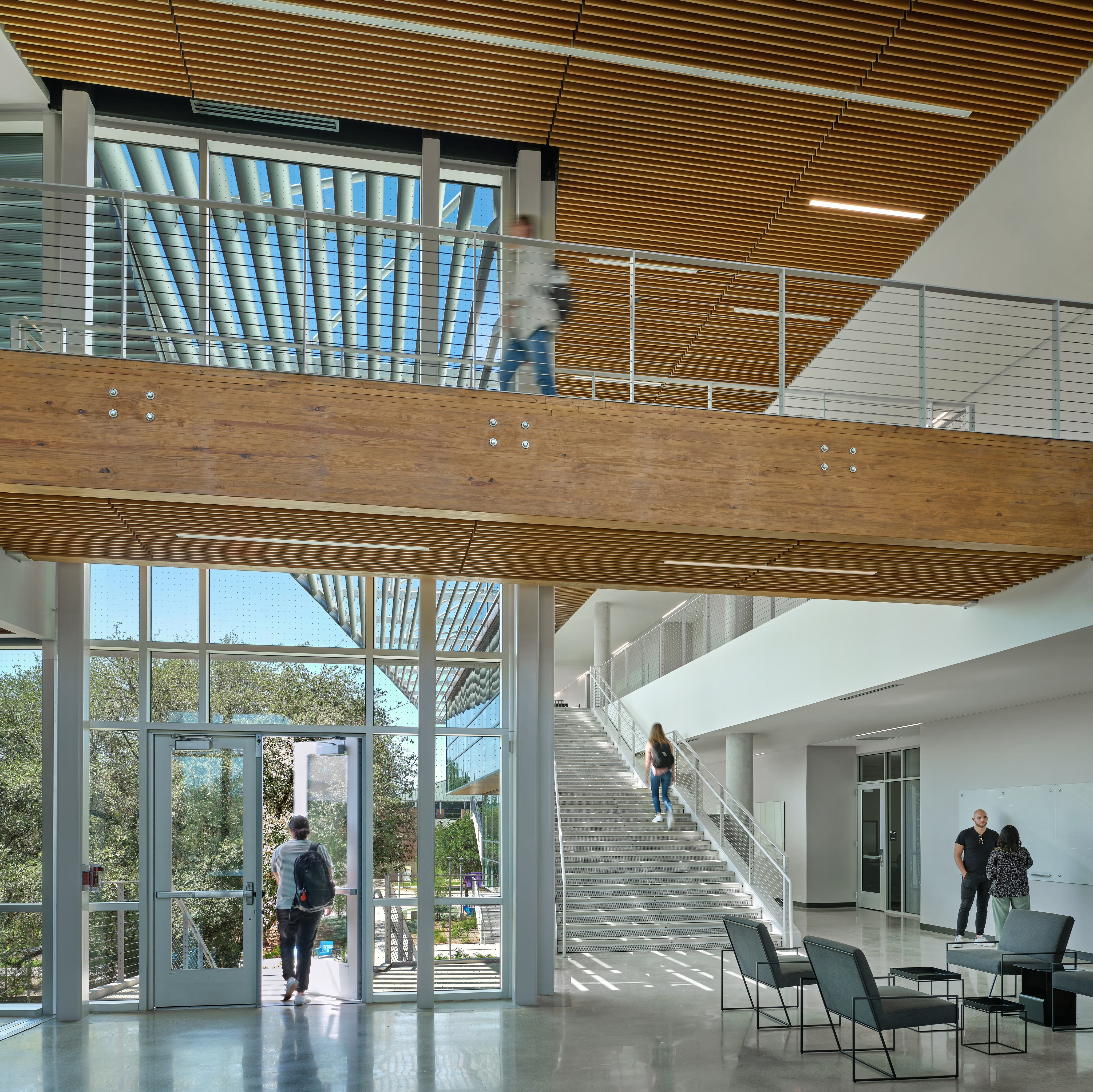 Modern building interior with a wooden mezzanine. Two people walk on the upper level, while three converse at the base of a staircase. Another person ascends the stairs. The Cedar Elm STEM Center showcases glass and metal architecture throughout, embodying the spirit of Northwest Vista College.