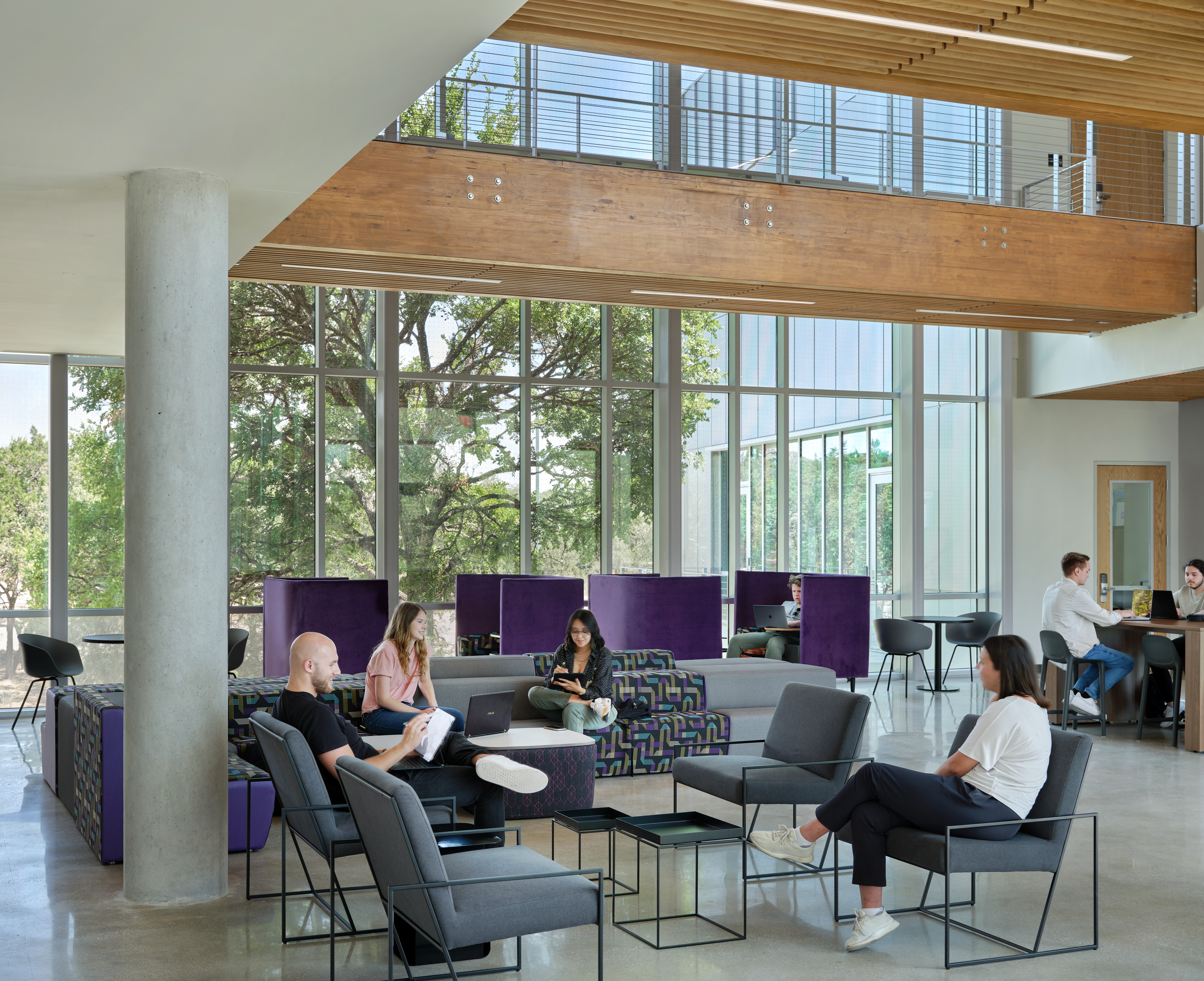Modern office space at the Northwest Vista College STEM Center with people seated on various chairs and sofas, working on laptops or talking. Large windows and open layout with natural light. Elevated walkway visible in the background.