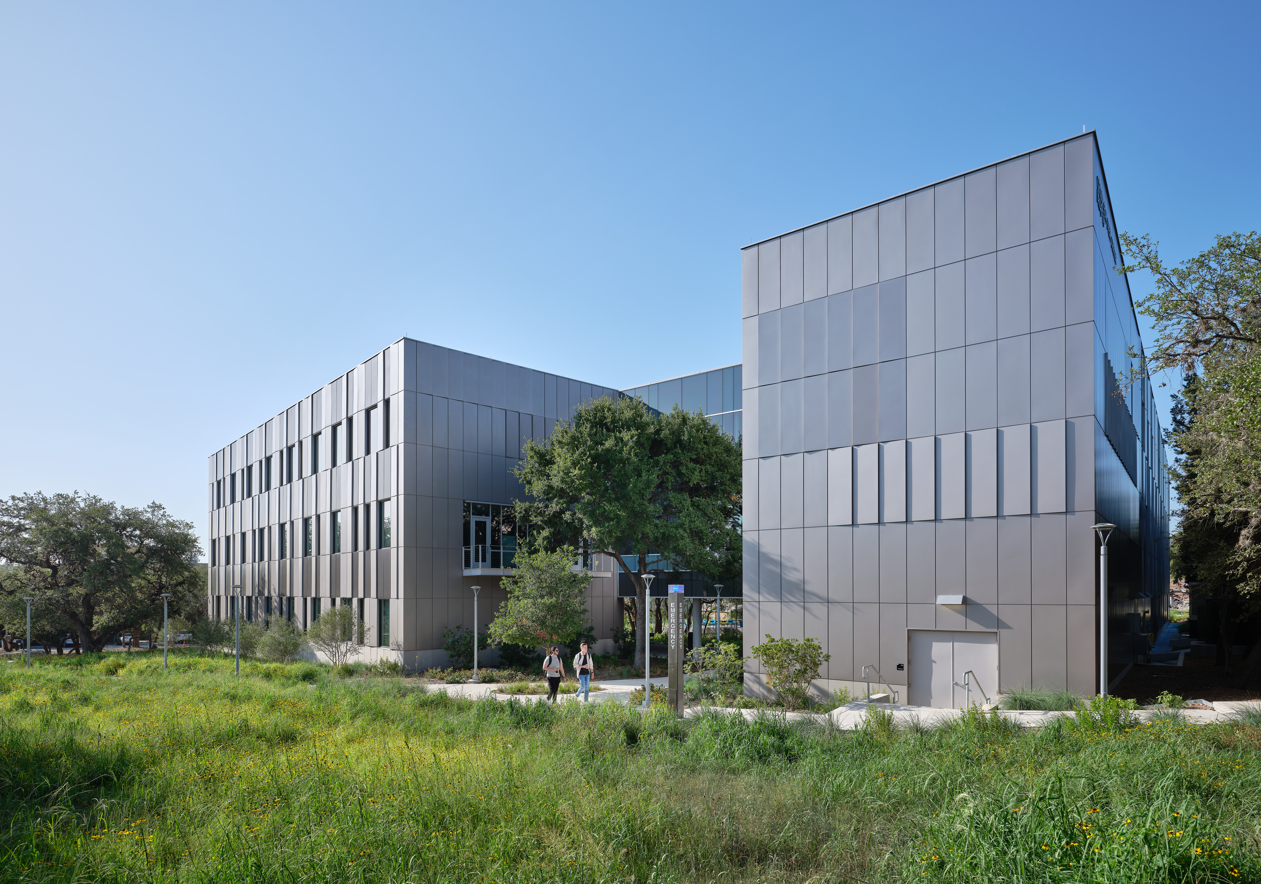 The modern three-story building with a sleek, metallic exterior, surrounded by greenery and trees under a clear blue sky, stands proudly as the new STEM Center at Northwest Vista College. Two people walk on a path near the entrance, embodying the vibrant life of Alamo Colleges.
