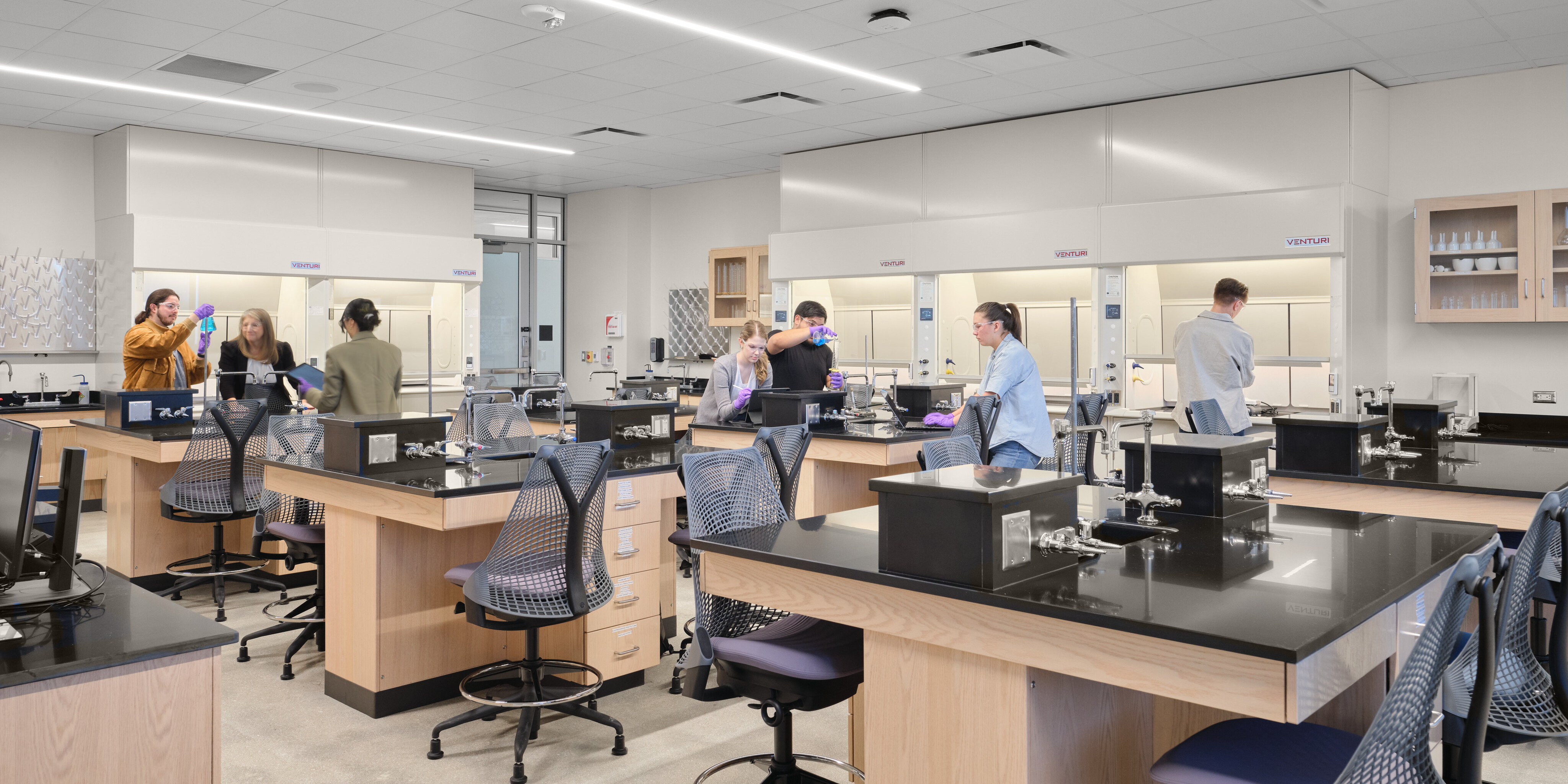 A group of people work in a modern laboratory with wooden benches, black countertops, and high chairs at the STEM Center in Northwest Vista College. Lab equipment and storage cabinets are visible in the background, showcasing the state-of-the-art facilities provided by Alamo Colleges.