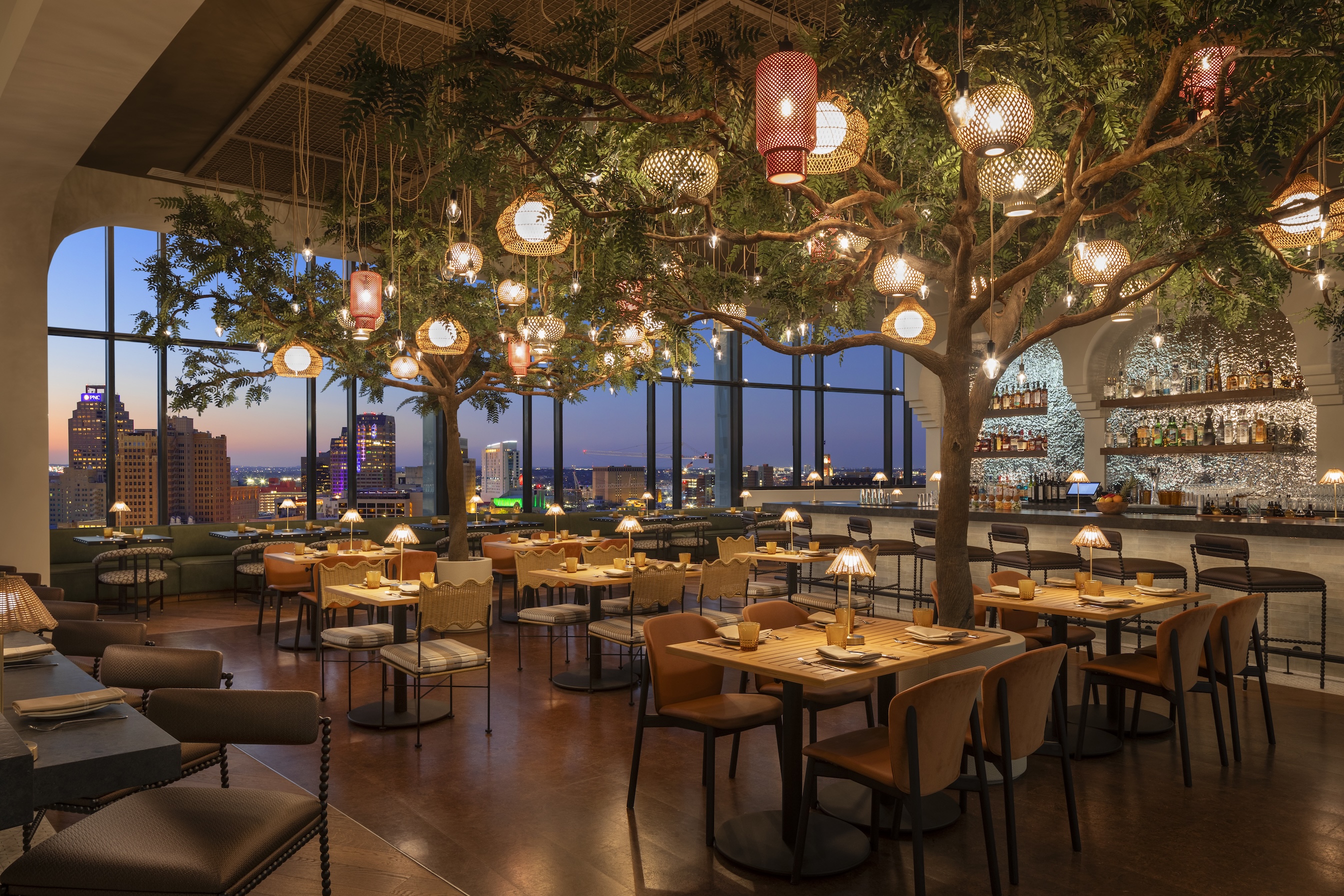 Upscale restaurant at Monarch San Antonio with indoor trees, hanging lanterns, and city skyline views through large windows at sunset; tables are set for dining, and a lit bar glows in the background of this Curio Collection Hilton San Antonio hotel.