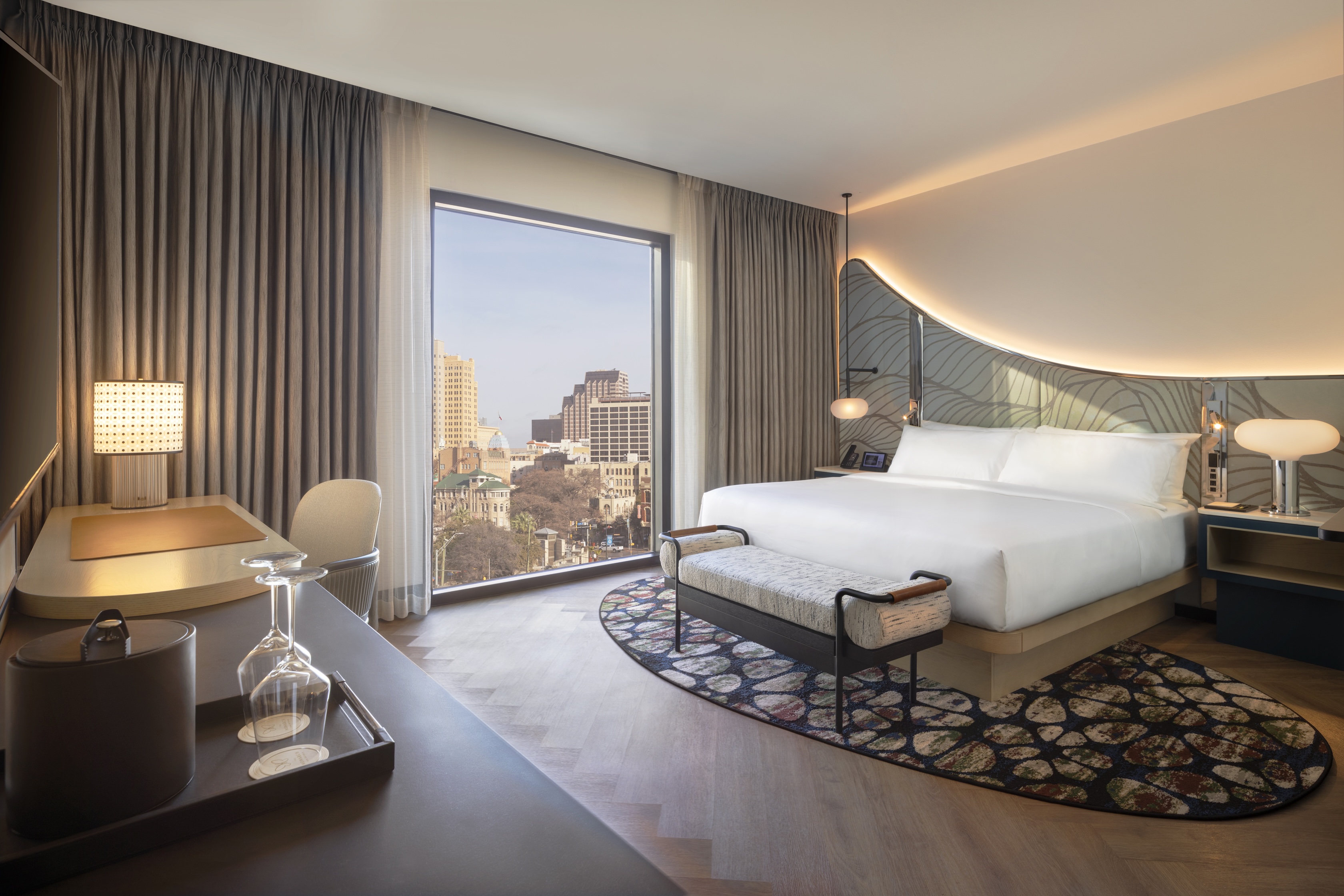 Modern hotel room at Monarch San Antonio, Curio Collection by Hilton, featuring a large bed, desk, chair, stylish lighting, cityscape views through the window, and contemporary decor in this upscale San Antonio hotel.