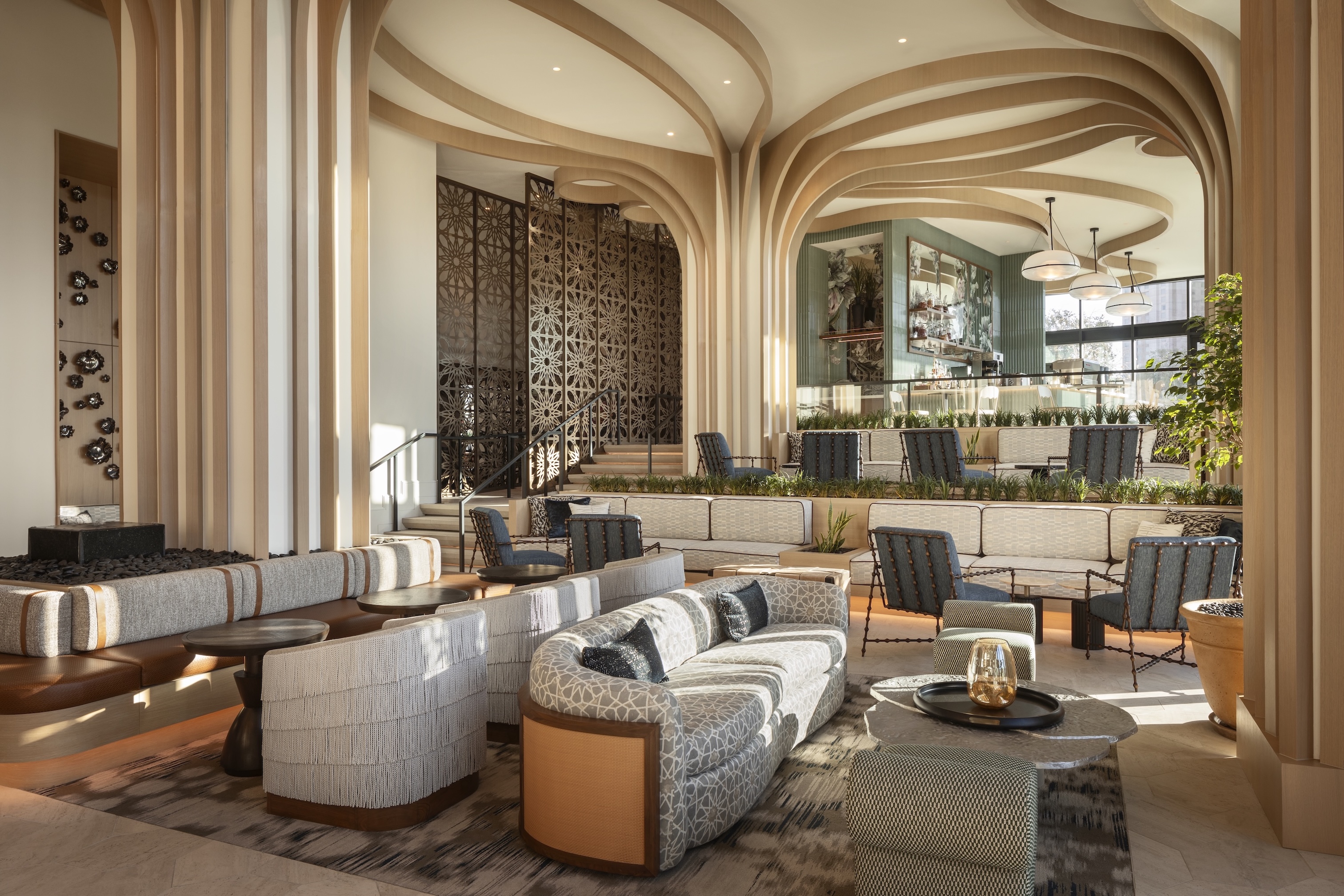 Modern lounge at Monarch San Antonio with curved wooden ceiling panels, patterned sofas, armchairs, and a bar area in the background—part of the Curio Collection by Hilton—featuring decorative screens and abundant natural light.