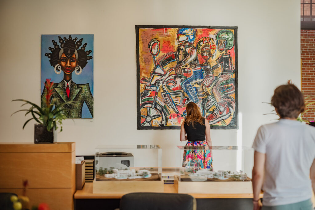 A woman stands facing a colorful painting by a San Antonio artist on a gallery wall, while another person observes from a distance; two art pieces and model displays inspired by Africa are visible.