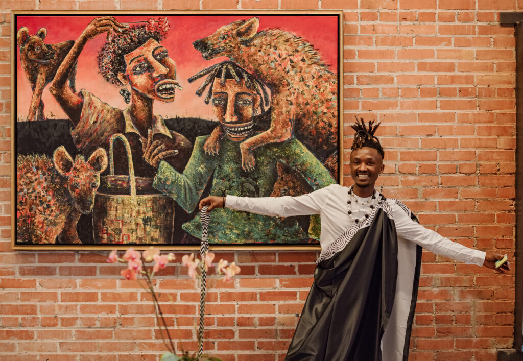 A person in black and white attire poses smiling in front of a colorful painting by a San Antonio artist, depicting two figures with hyenas, set against a brick wall—capturing the spirit of storytelling through art.