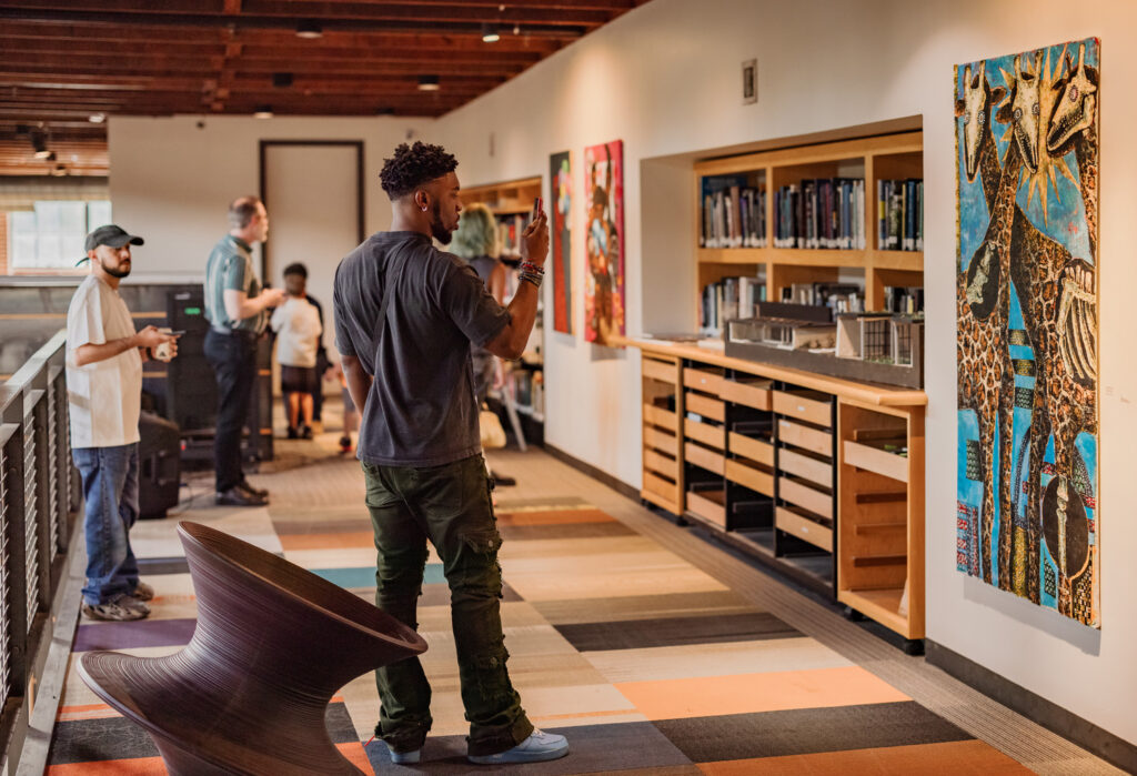 A man observes a colorful abstract painting by a San Antonio artist in a modern gallery space while others look around and interact in the background.