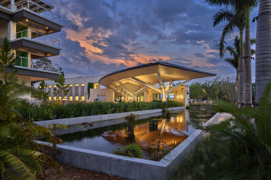 Modern building with a distinctive angular roof by Overland Partners, reflected in a pond and surrounded by lush greenery and palm trees at sunset in the Marie Selby Botanical Gardens, an AIA COTE Top Ten Award winner.