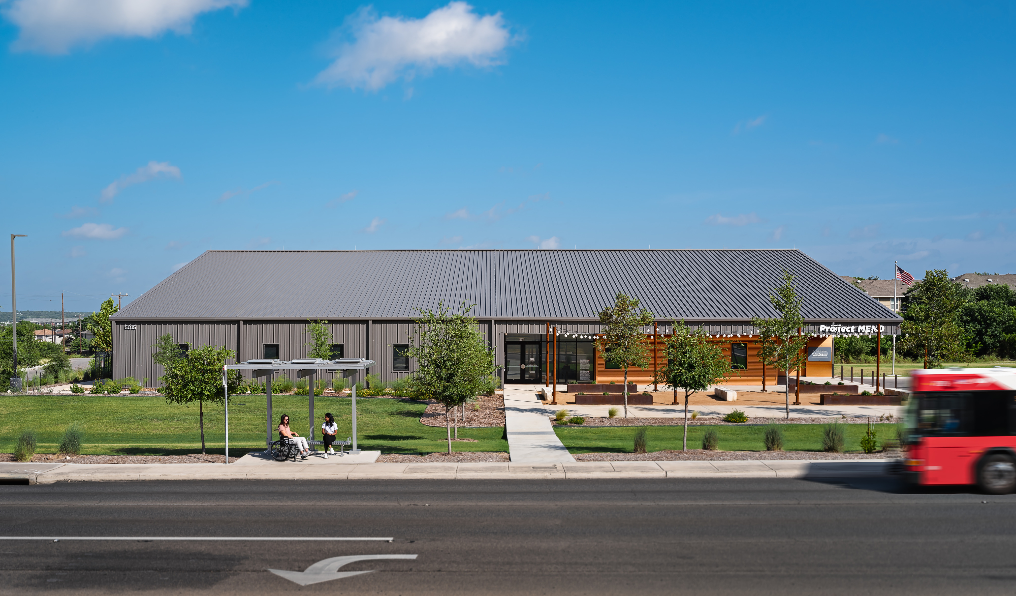 A gray metal building sits behind a lawn with trees; a Project MEND bus stop with three people and a passing red bus are in the foreground on a sunny day.