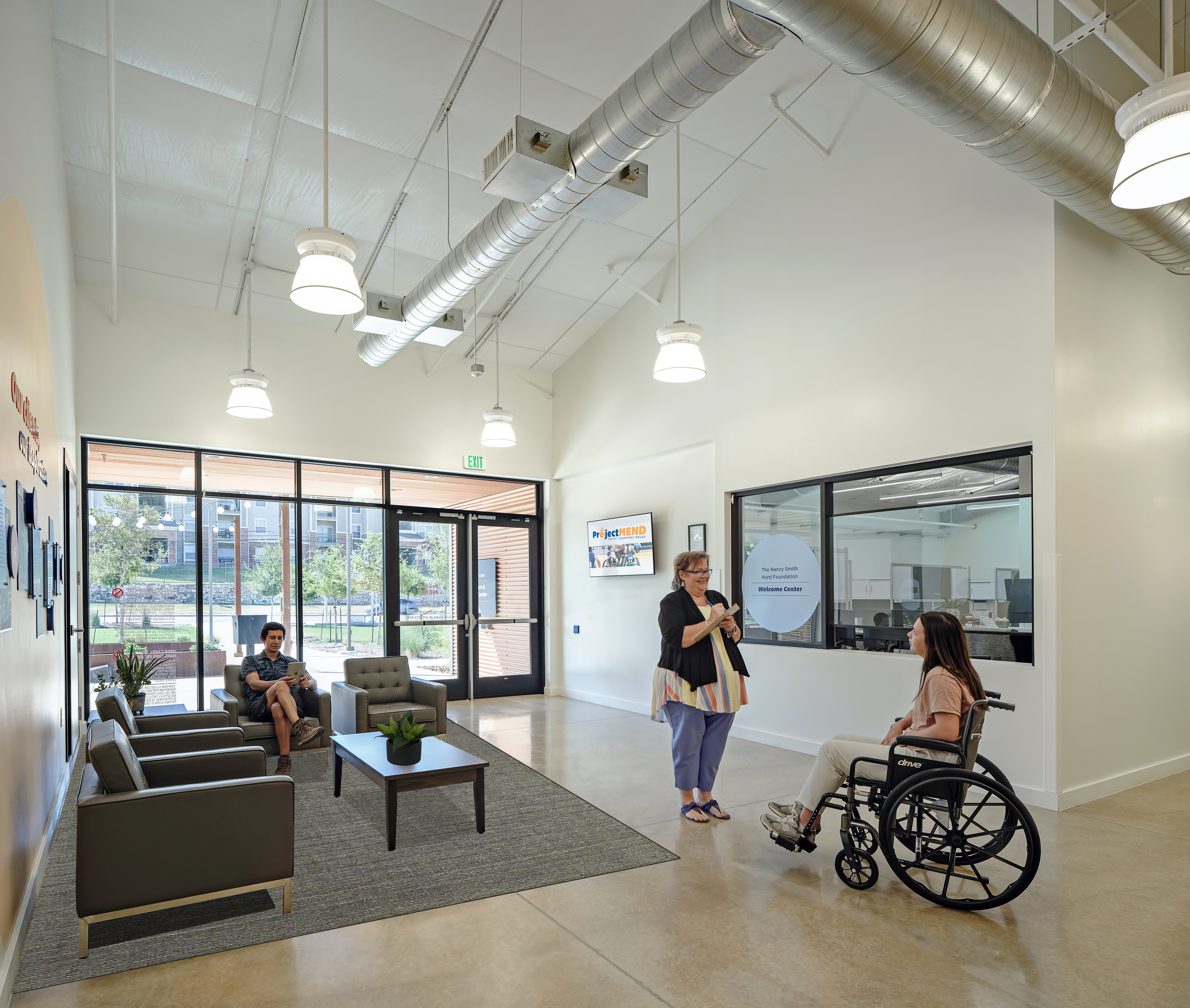 A person in a wheelchair and another individual are discussing Project MEND in a modern office lobby, while another person sits on a sofa using a phone; large windows provide natural light.