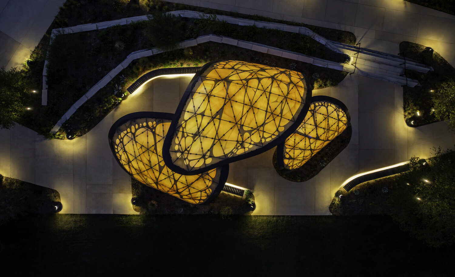 Aerial view of illuminated yellow domed structures with lattice patterns surrounded by walkways, landscaping, and soft ground lighting at night.