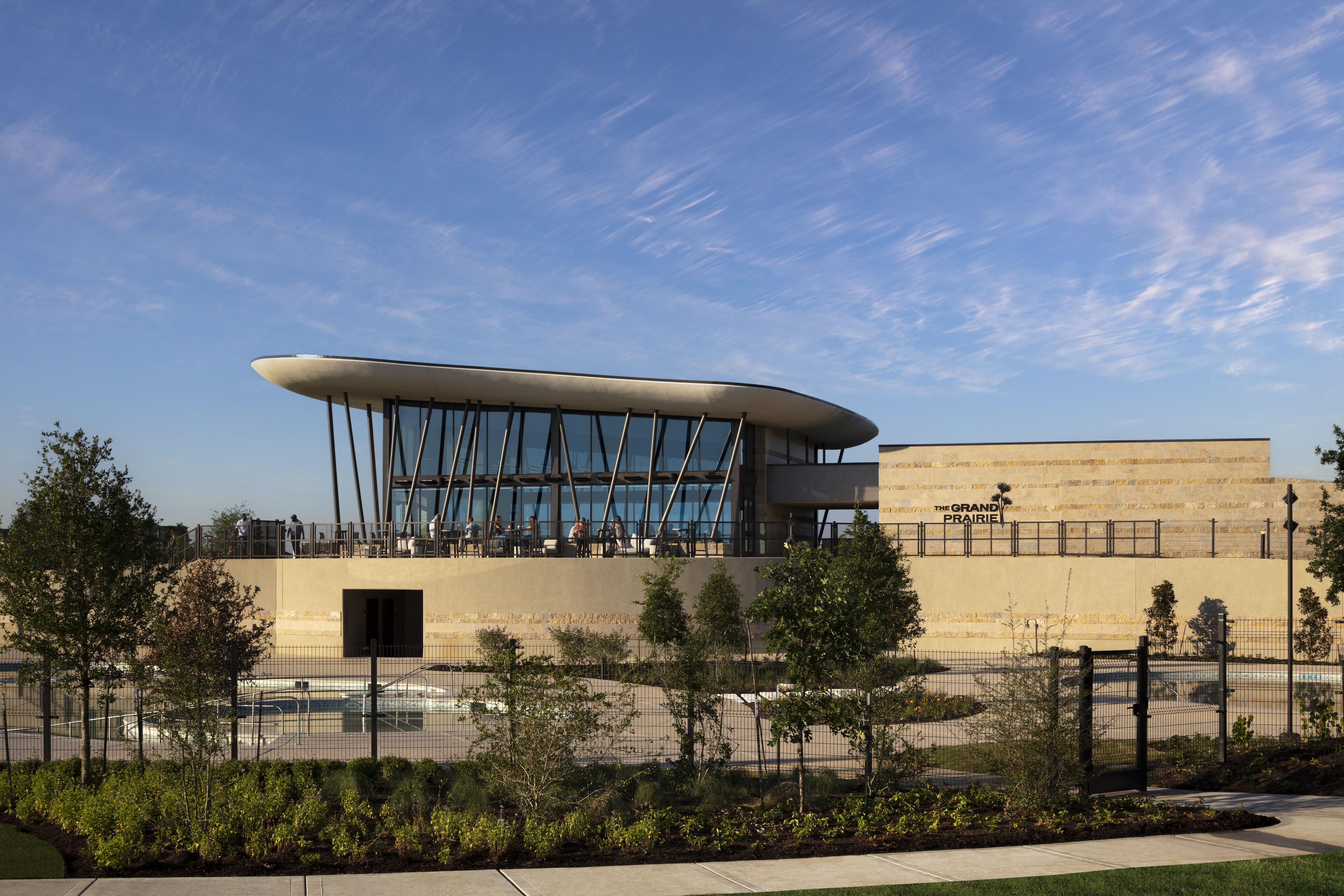 Modern beige building with large glass windows and a cantilevered roof, the Sundancer Amenity Complex is surrounded by landscaped greenery under a blue sky with wispy clouds.