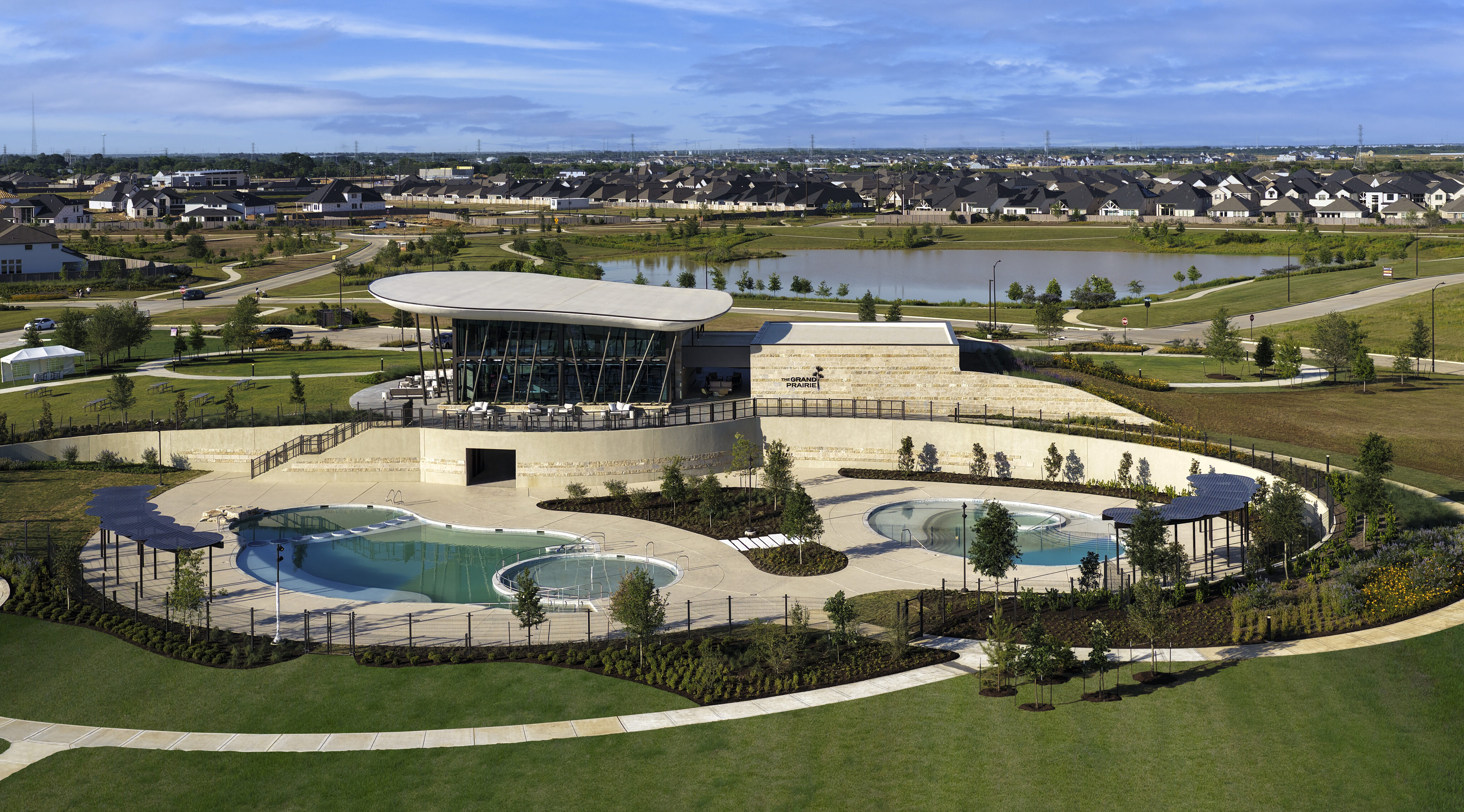Modern Amenity Complex with two outdoor swimming pools and landscaped grounds, surrounded by residential houses under a partly cloudy sky—a vibrant centerpiece of the Sundancer community.