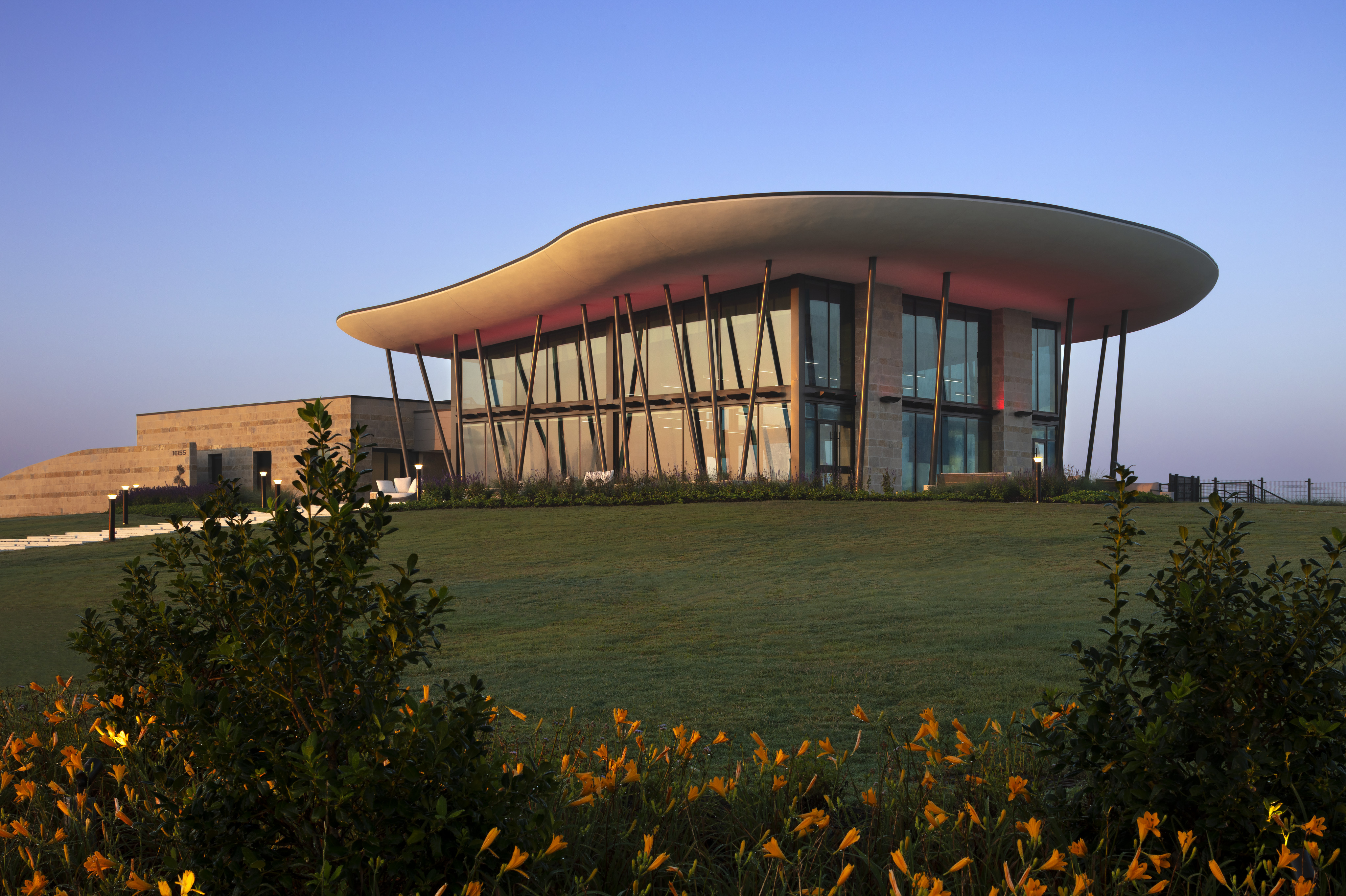 Modern building with a curved roof and tall windows, known as the Sundancer Amenity Complex, surrounded by green lawn and yellow flowers, under a clear sky.